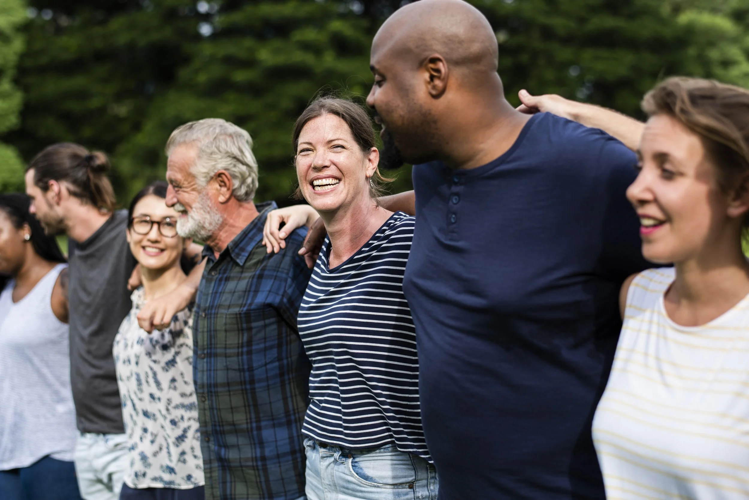 Group of diverse people standing outdoors with arms around each other's shoulders, smiling and enjoying a sunny day