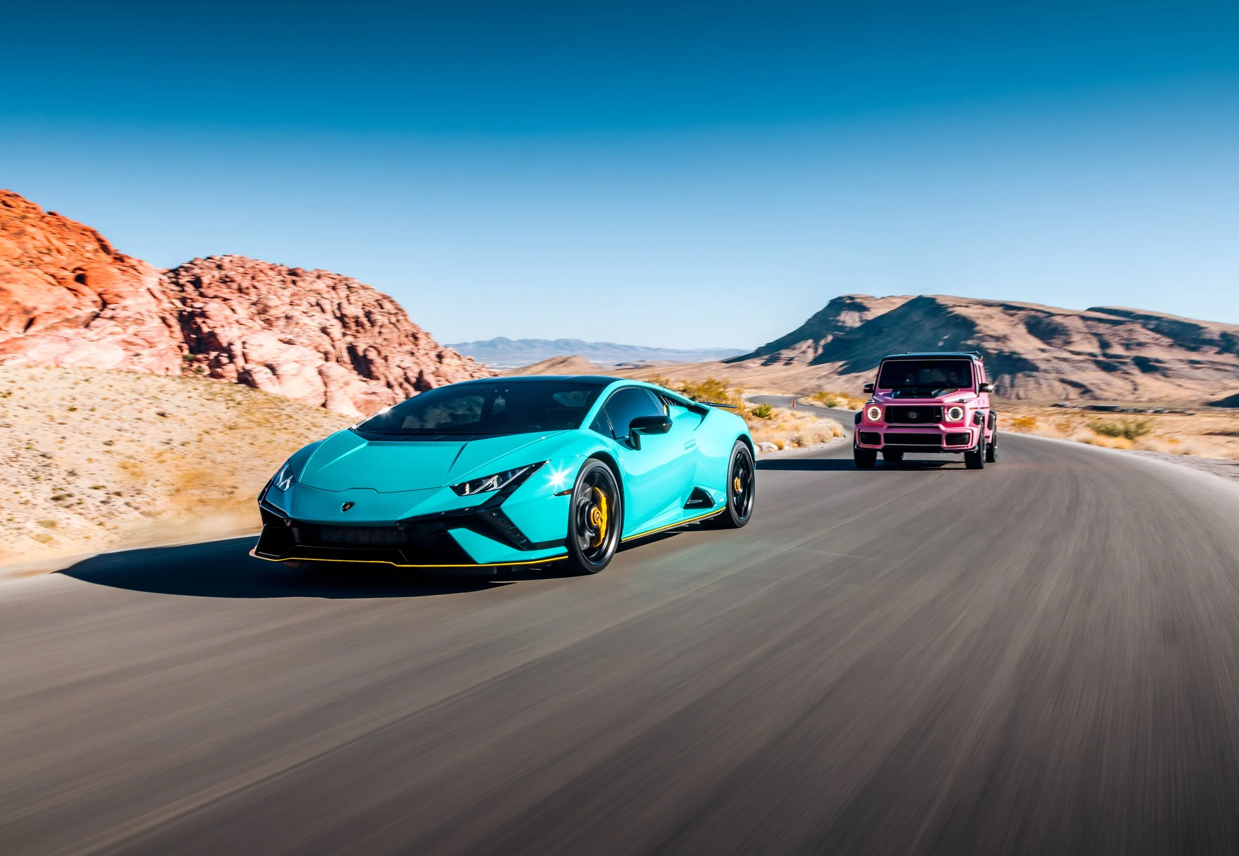 A blue Lamborghini sports car and a pink SUV driving on a desert highway with rocky hills and mountains in the background under a clear blue sky.