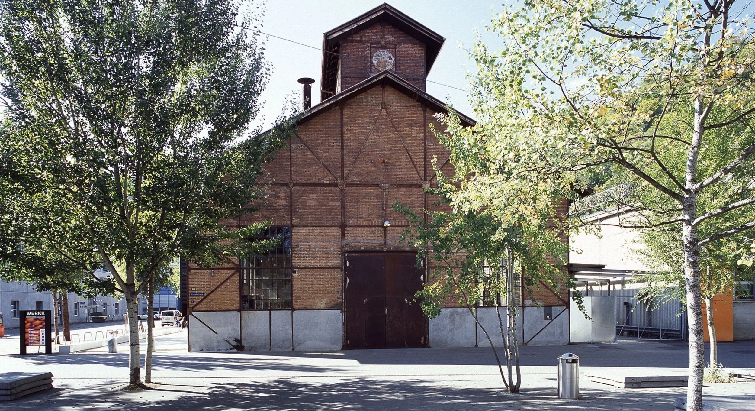 An old brick building with a small tower on top, surrounded by trees and benches in a park-like setting.