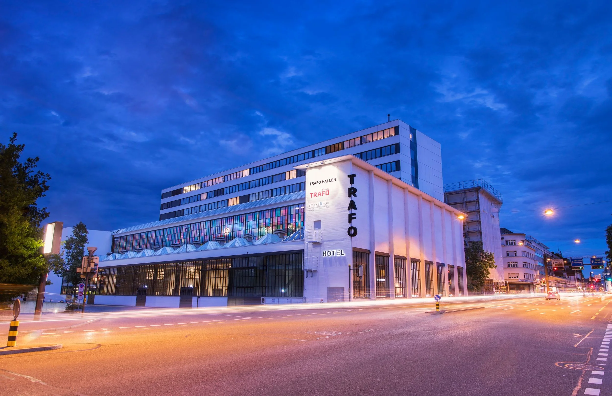 Night view of the Trafo Hotel building with illuminated signage and colorful interior lights, street with moving vehicle light streaks, and a dark blue sky with clouds.