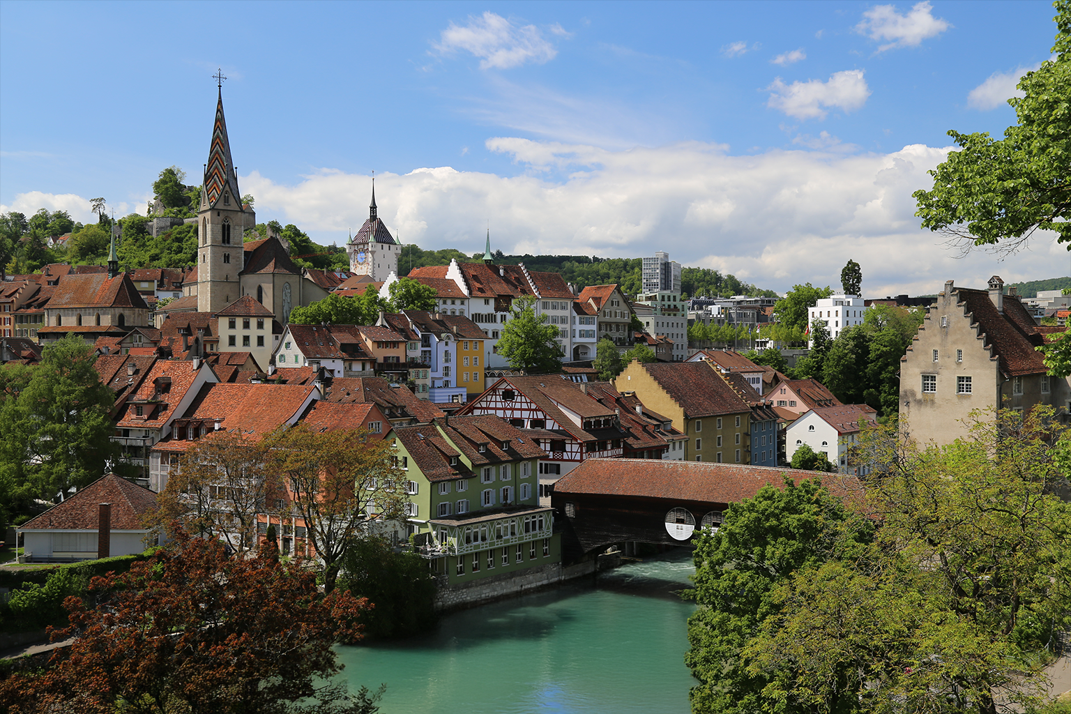 A scenic view of a European town with colorful buildings, red-tiled roofs, a church with a tall steeple, and a river flowing through the town, surrounded by green trees and hills under a partly cloudy sky.