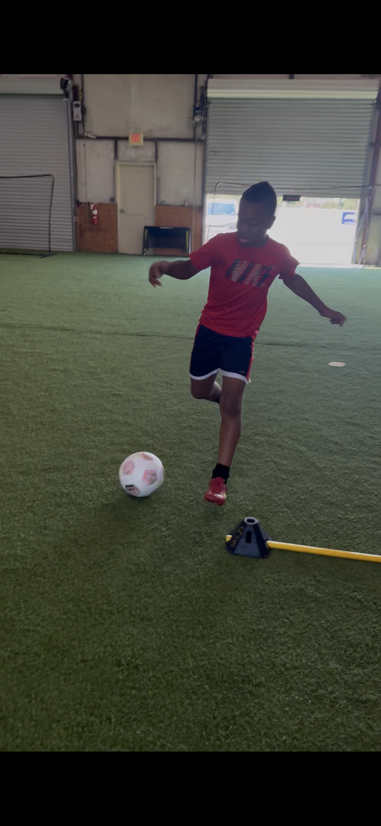 A young boy in a red shirt and black shorts is practicing soccer indoors on a green artificial turf field, kicking a white soccer ball with pink and black accents.