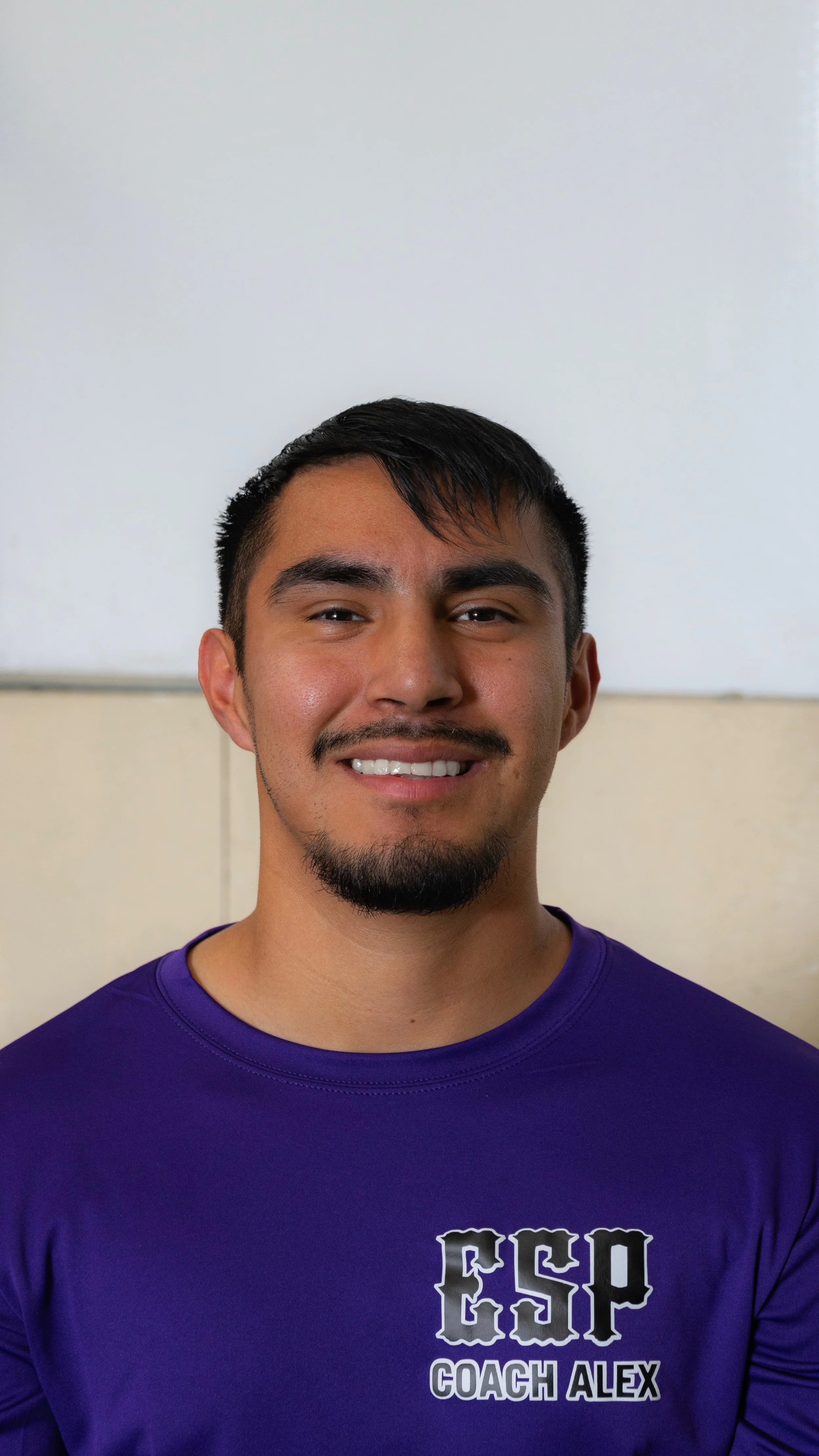A young man with dark hair, a goatee, and a mustache smiling, wearing a purple shirt with the text 'CSP Coach Alex' printed on it, standing in front of a light-colored wall.