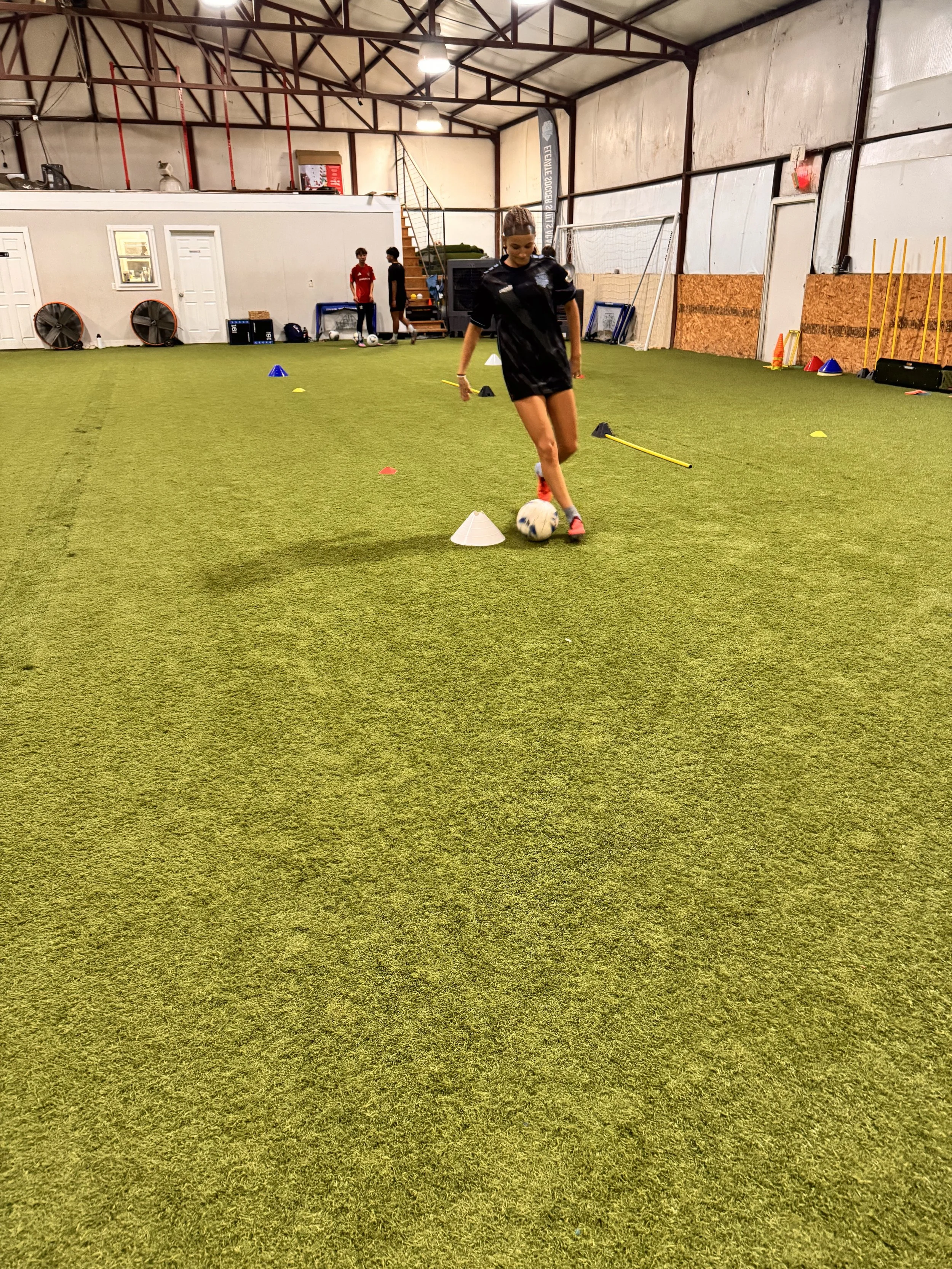 A young woman in black training gear practicing soccer skills indoors, maneuvering a soccer ball around cones and agility poles marked with different colors on artificial turf.