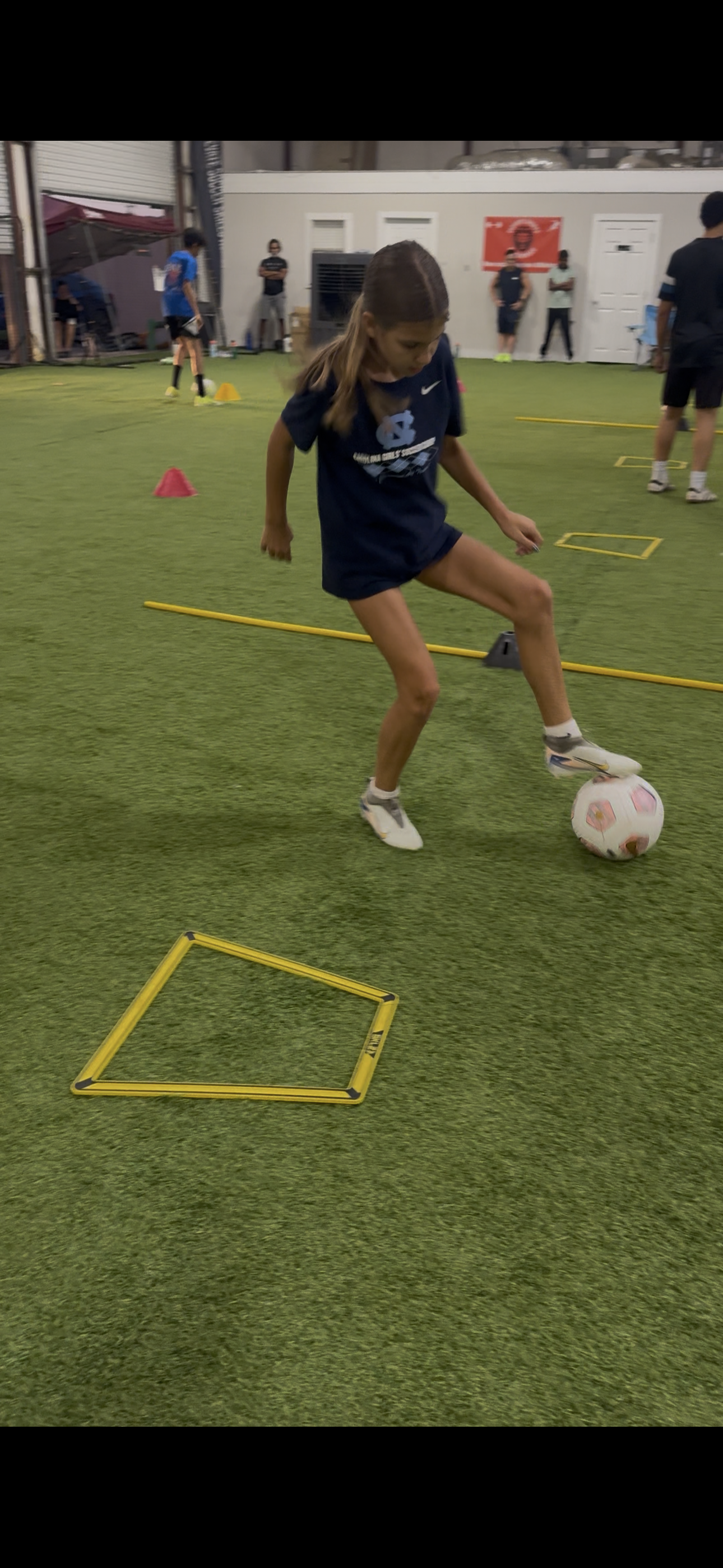 A young girl in a navy sports outfit practicing soccer inside an indoor sports facility with artificial turf, yellow agility markers, and pink cones.