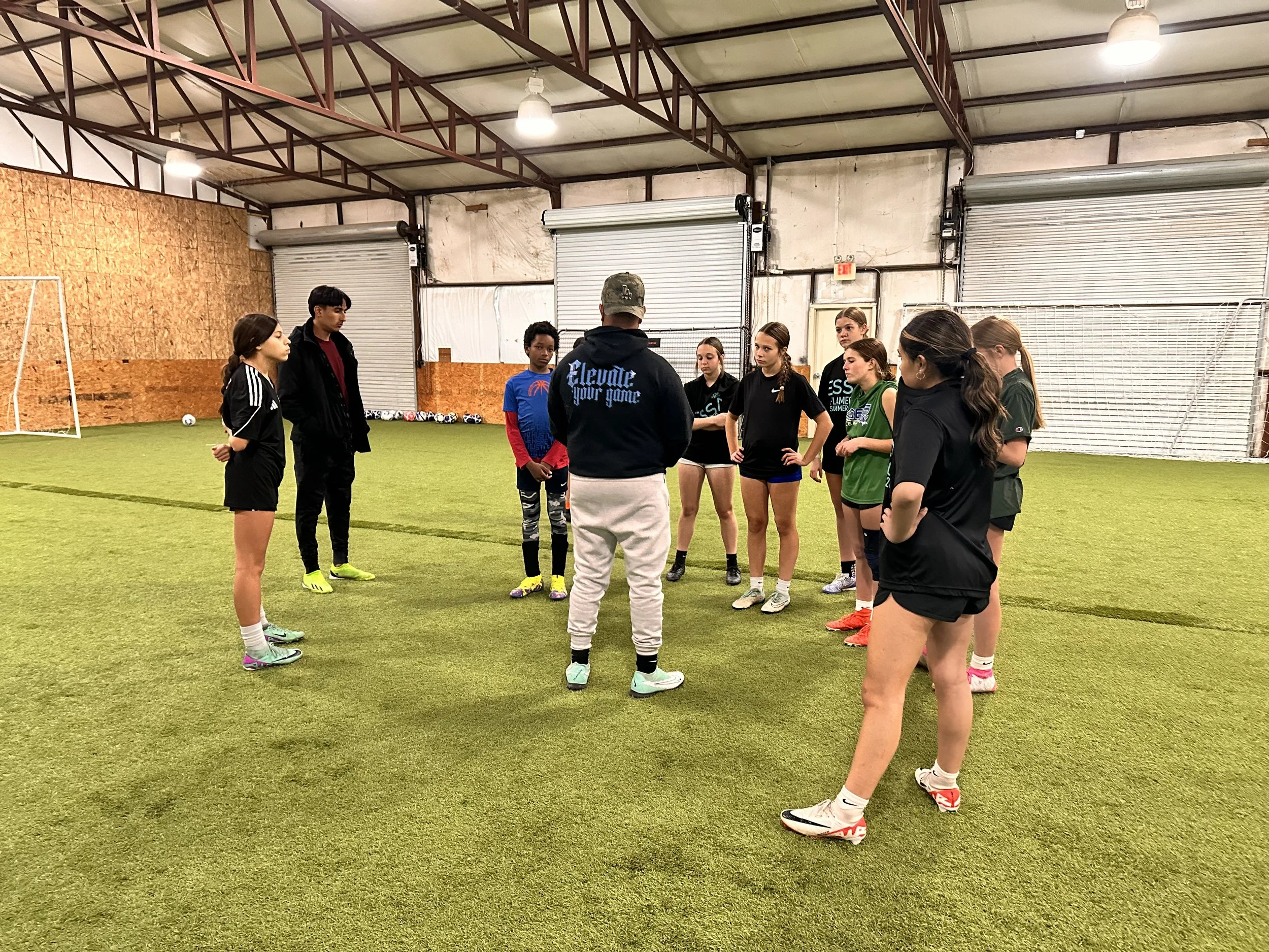 A group of young female soccer players and one male coach standing on an indoor artificial turf field, participating in a team discussion or receiving instructions. The players are dressed in sportswear, and the background shows a soccer goal and storage area with balls.