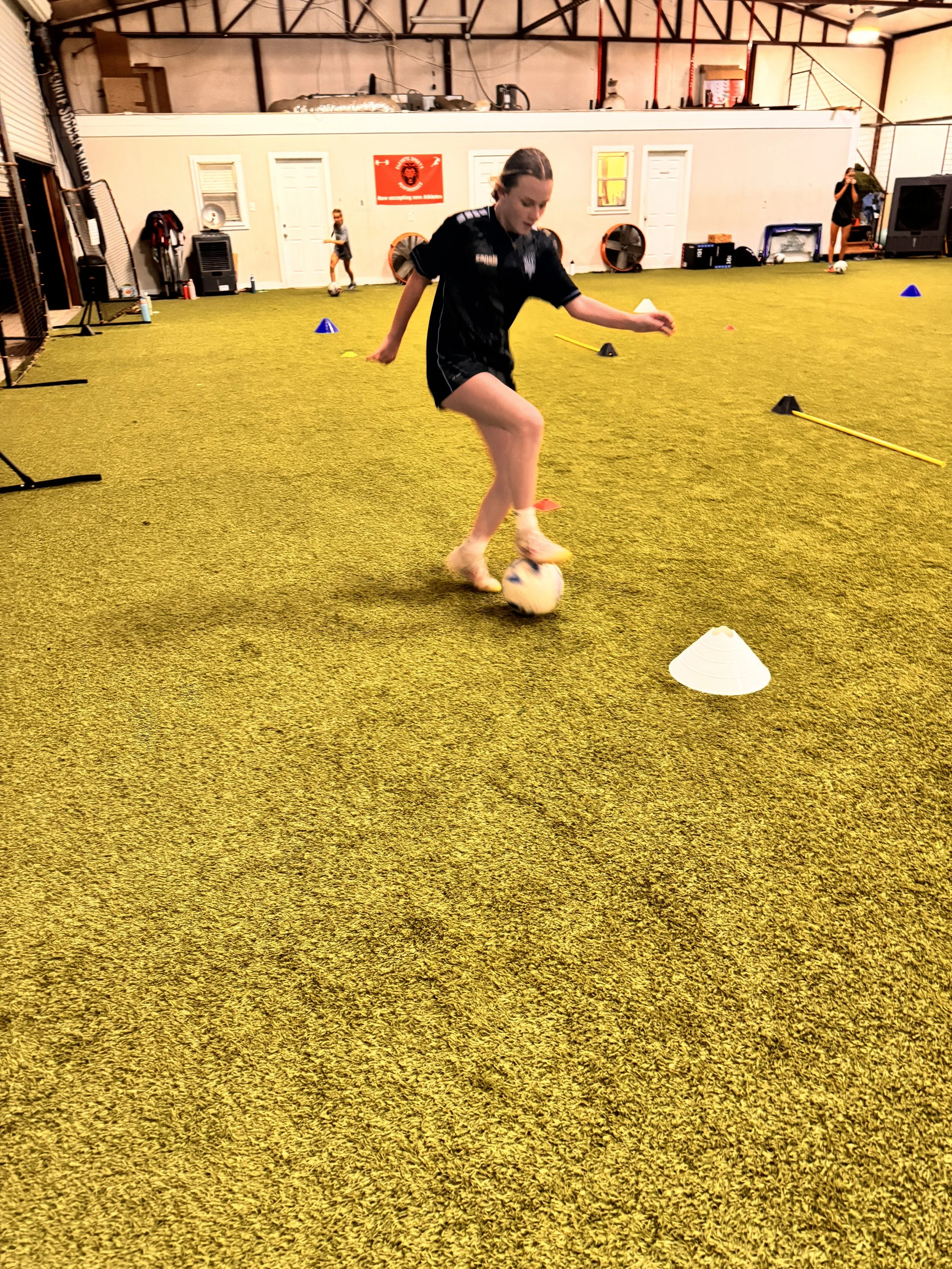 Young girl practicing soccer dribbling drills on an indoor turf field, surrounded by cones and training equipment.