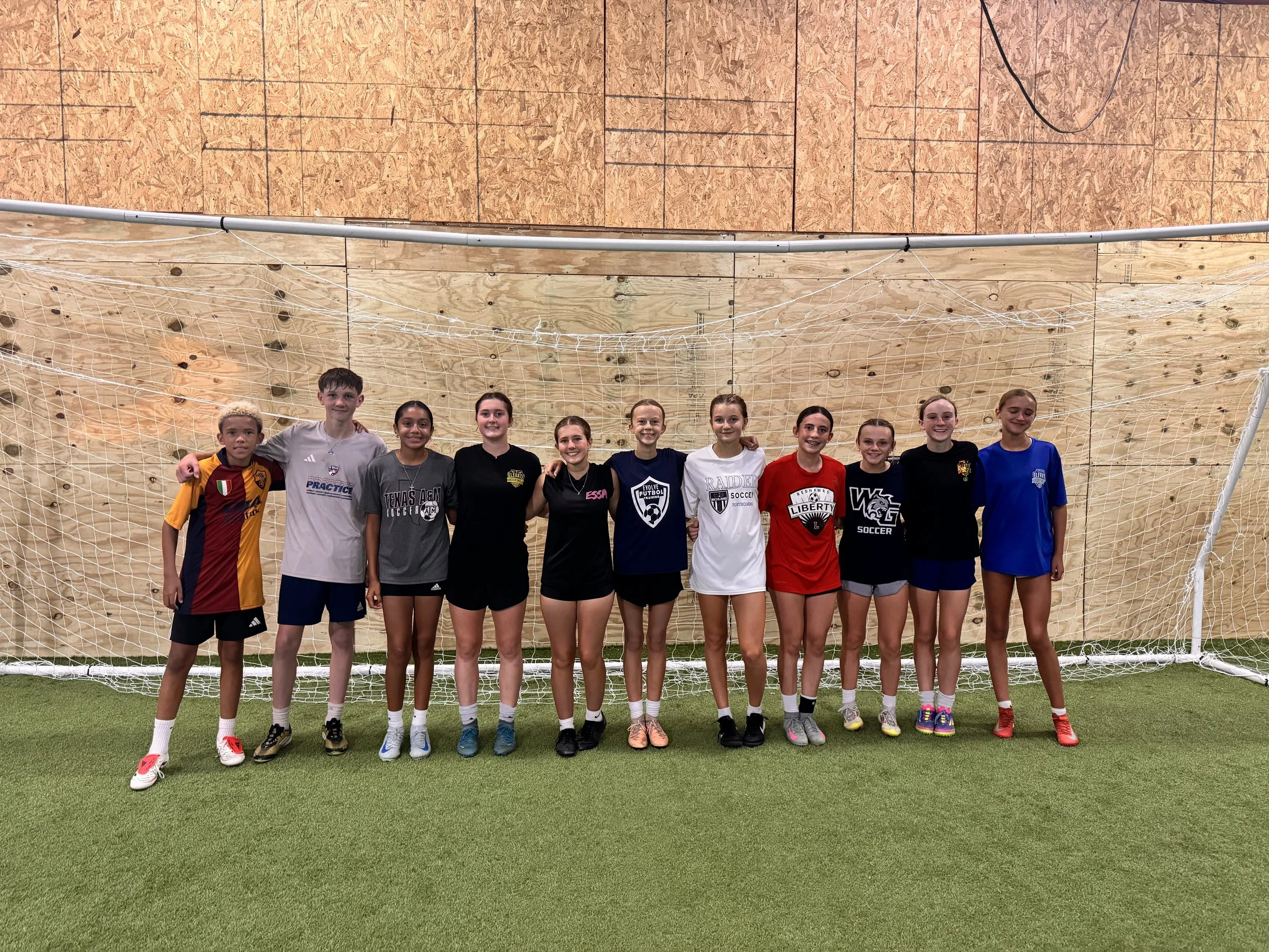 A group of eleven young girls standing together on an indoor soccer field in front of a soccer goal net, smiling at the camera. They are wearing sports jerseys and shorts, with some in skate shoes, and behind them there is a wooden wall.