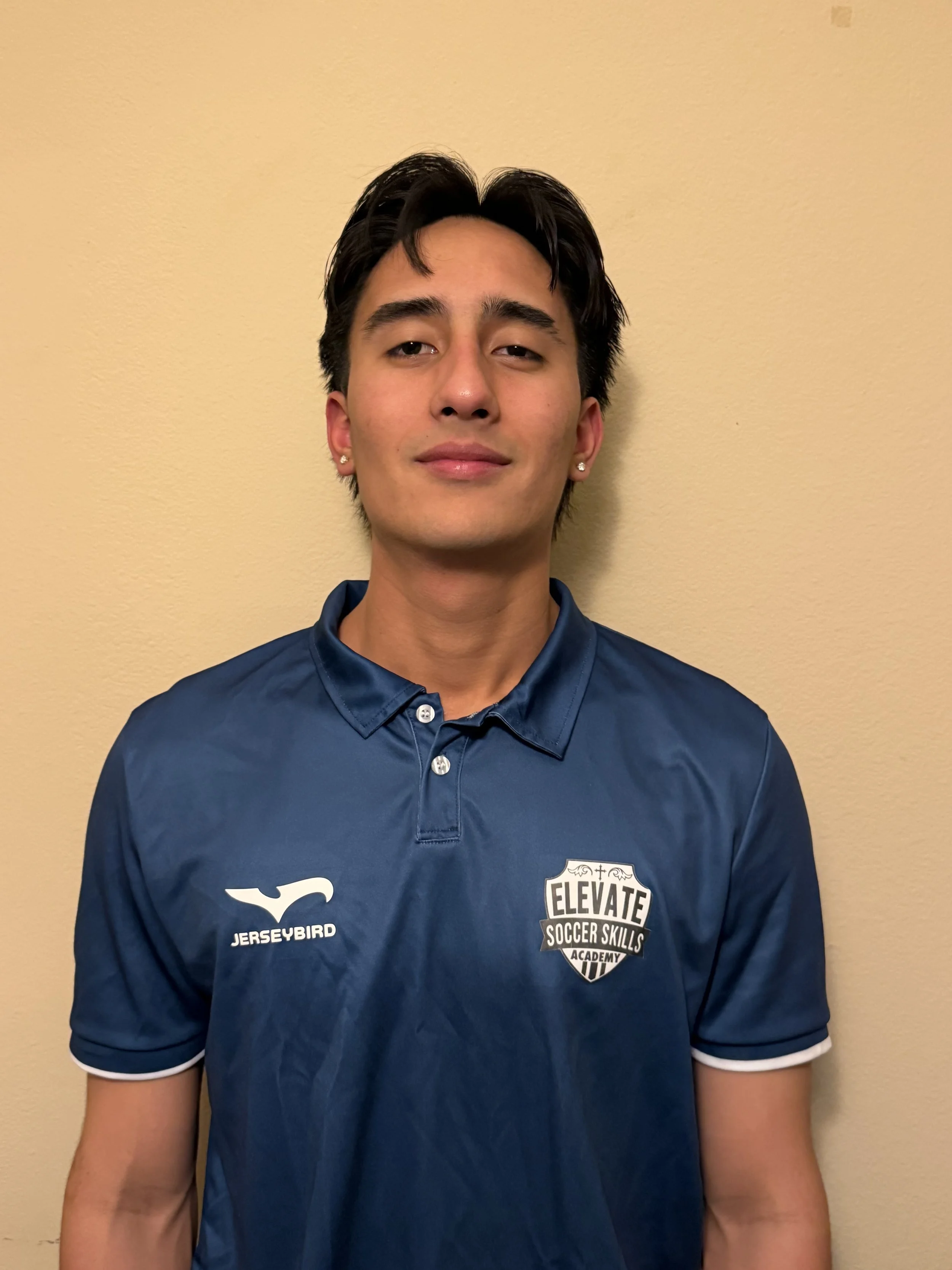 Young man with dark hair and earrings, wearing a blue soccer jersey with 'JERSEYBIRD' and 'ELEVATE SOCCER SKILLS ACADEMY' logos, standing against a beige wall.
