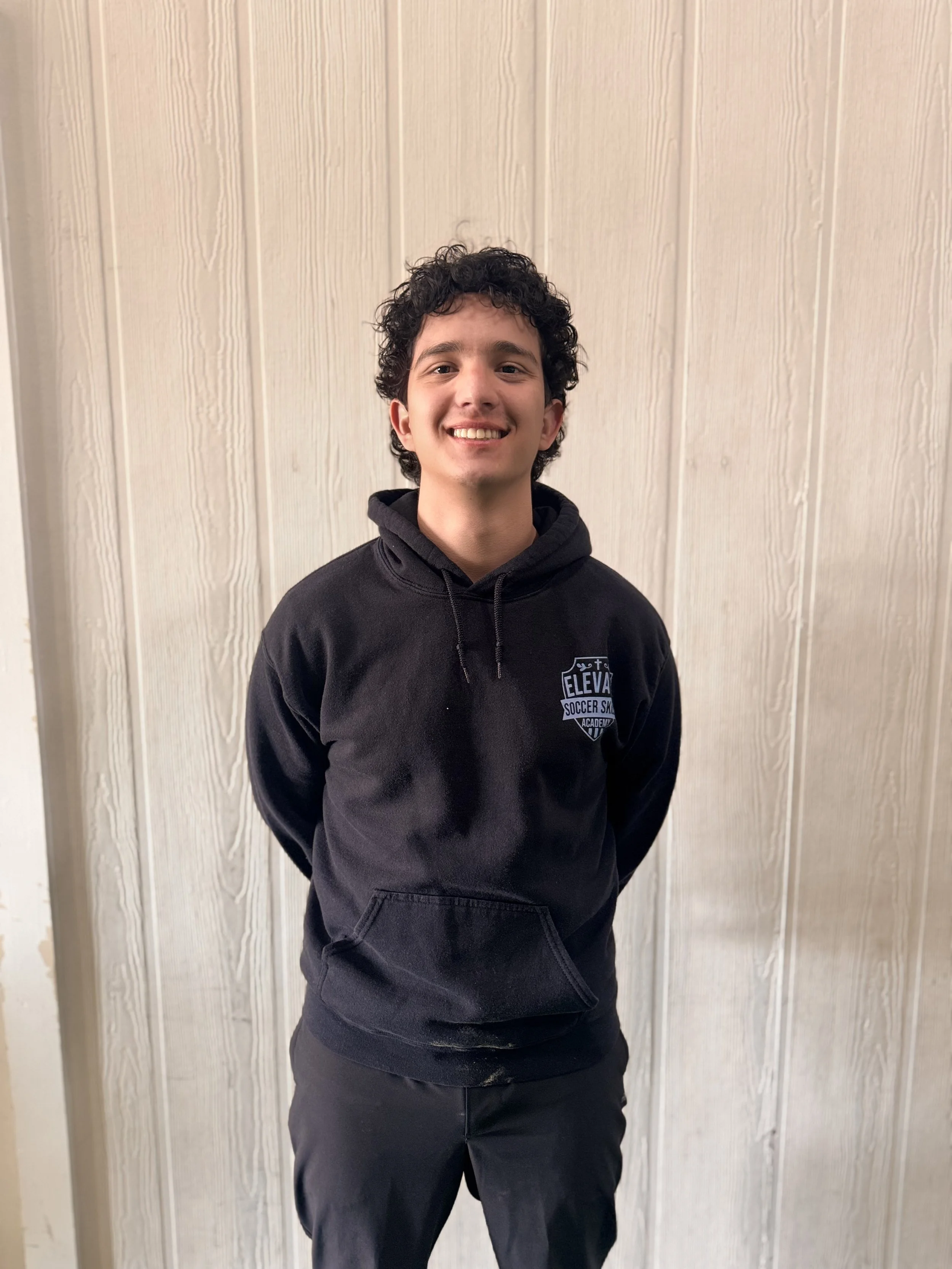 A young man with curly black hair smiling, wearing a black sweatshirt with a logo that says 'Elevation Soccer Skills Academy' and black pants, standing in front of a light-colored wooden wall.