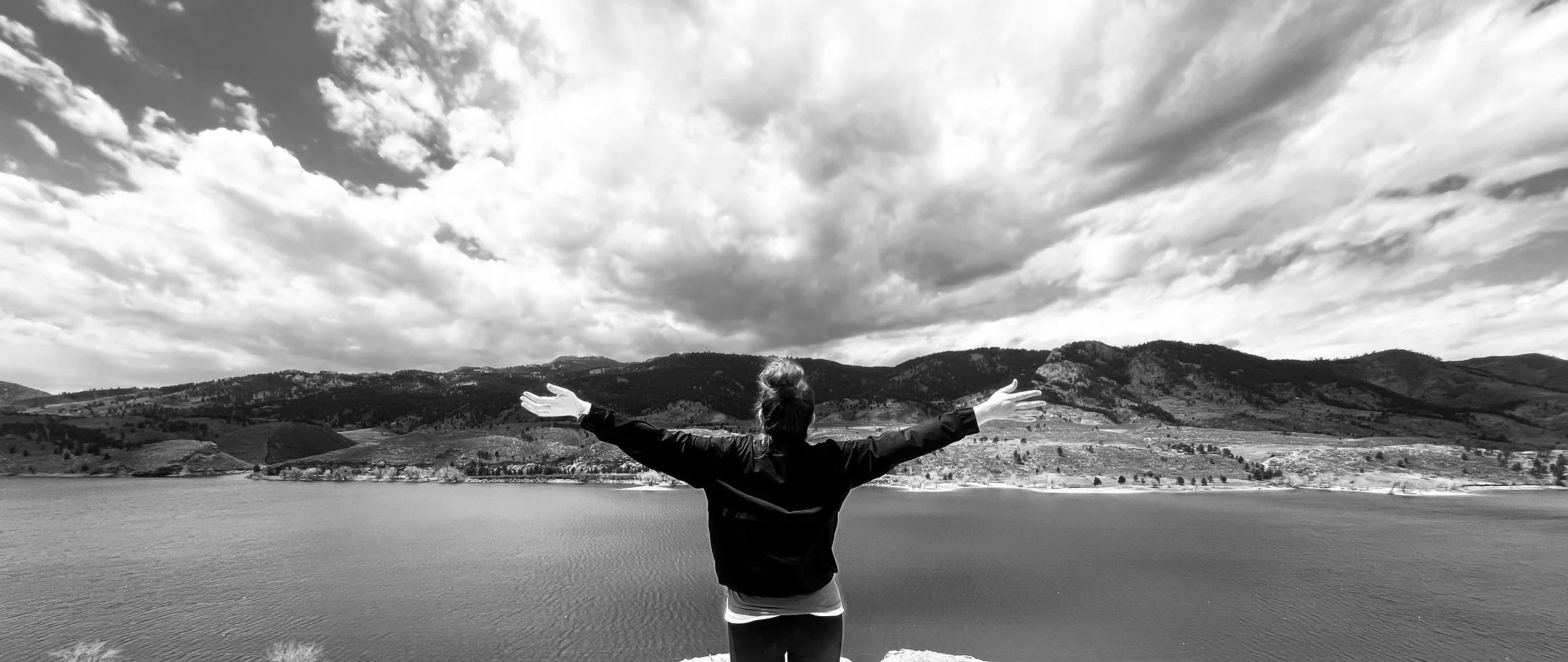 A person standing on a lakeshore with arms outstretched, facing a body of water and distant mountains under a cloudy sky.