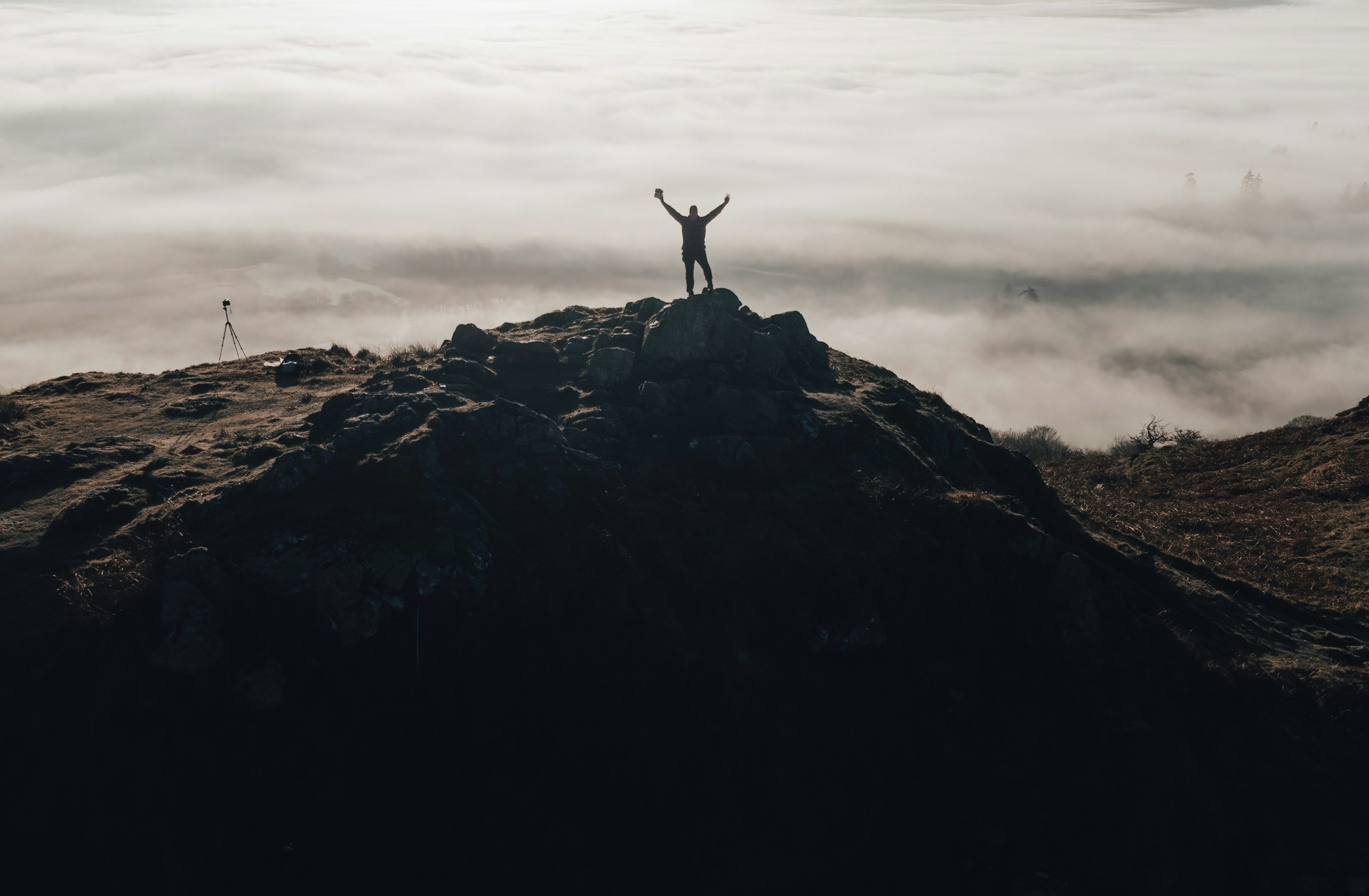 Person standing with arms raised on mountain peak, overlooking a foggy landscape with clouds below.
