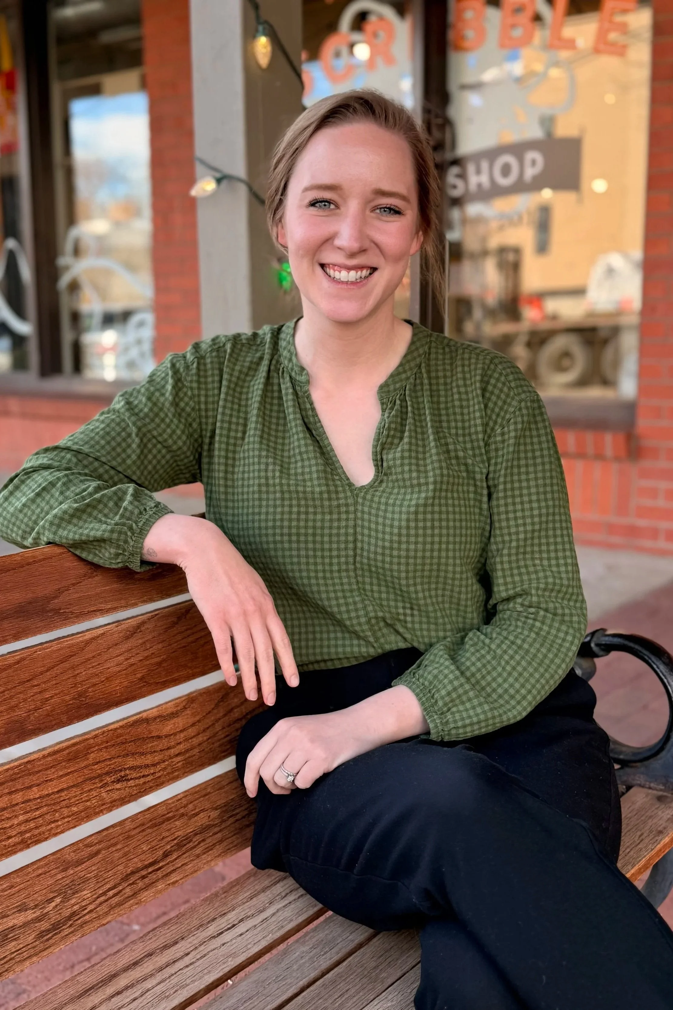 A woman sitting on a wooden bench outside a shop, smiling at the camera, wearing a green checkered blouse and black pants.