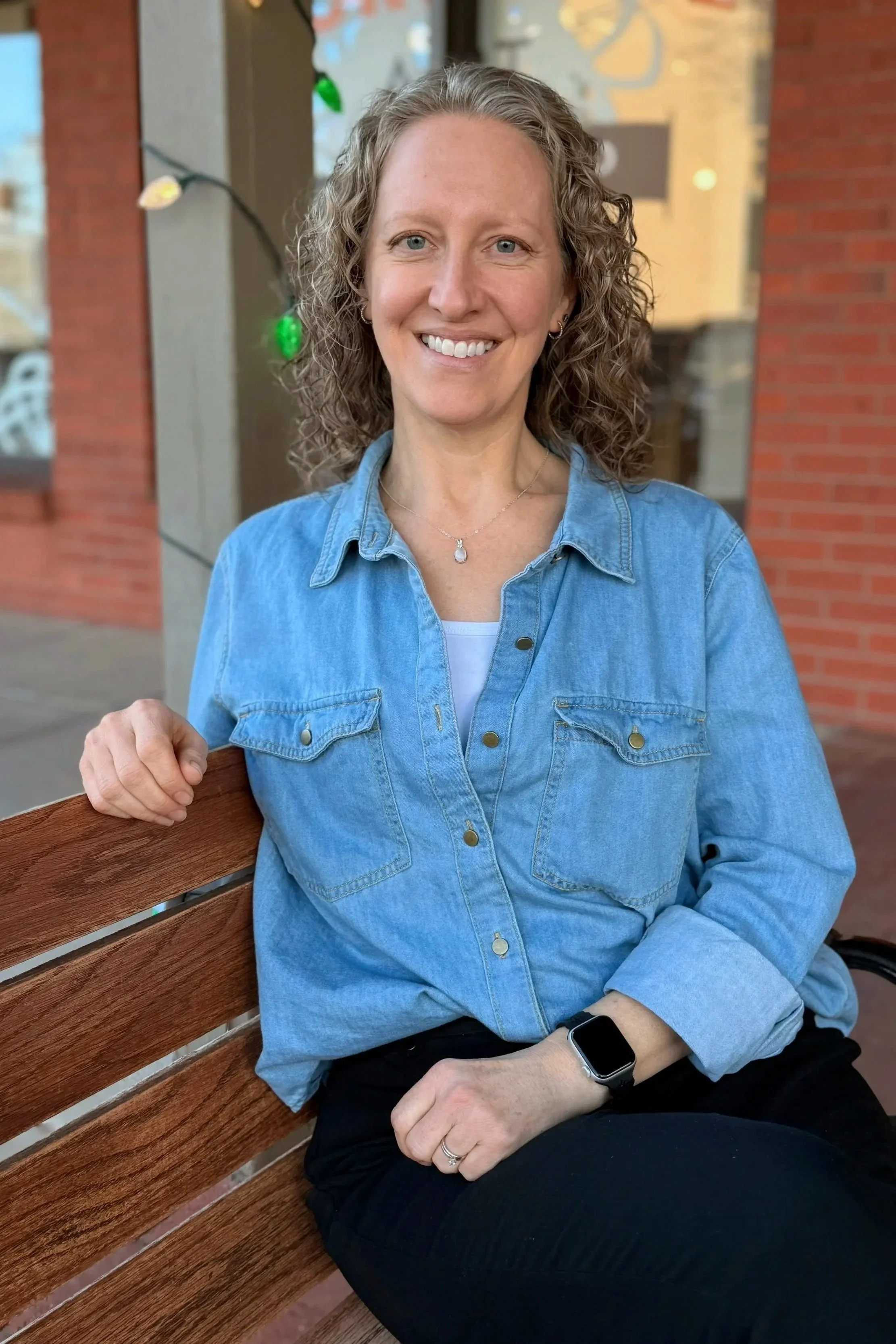 A woman with curly hair, wearing a denim shirt and black pants, sitting on a wooden bench outdoors, smiling at the camera with a blurred background of a building and some decorative lights.