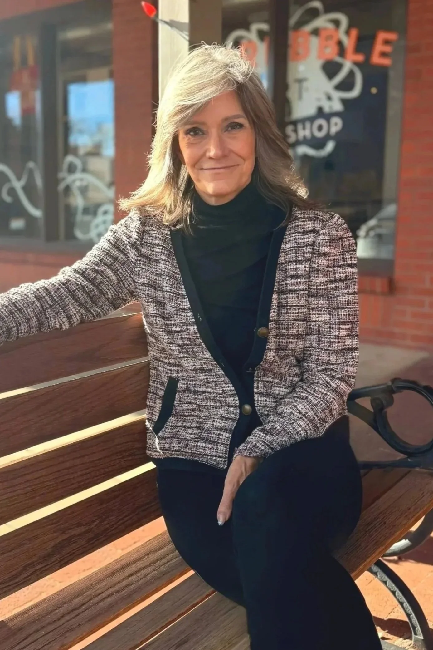 A woman with shoulder-length gray hair sitting on a wooden park bench outside a brick building with large windows. She is wearing a patterned blazer, black top, and black pants, smiling at the camera.