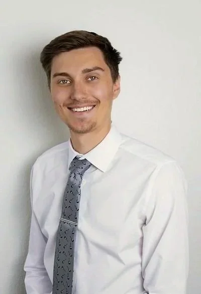 A man with short dark hair, light skin, and brown eyes is smiling slightly at the camera. He is wearing a collared shirt, with a plain blue background.