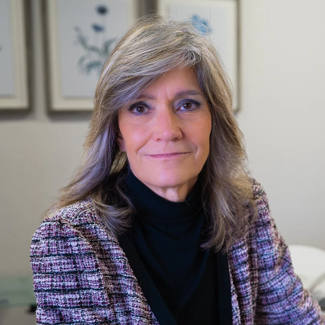 A woman with dark curly hair, wearing glasses, hoop earrings, a striped blouse, and layered necklaces, smiling at the camera.
