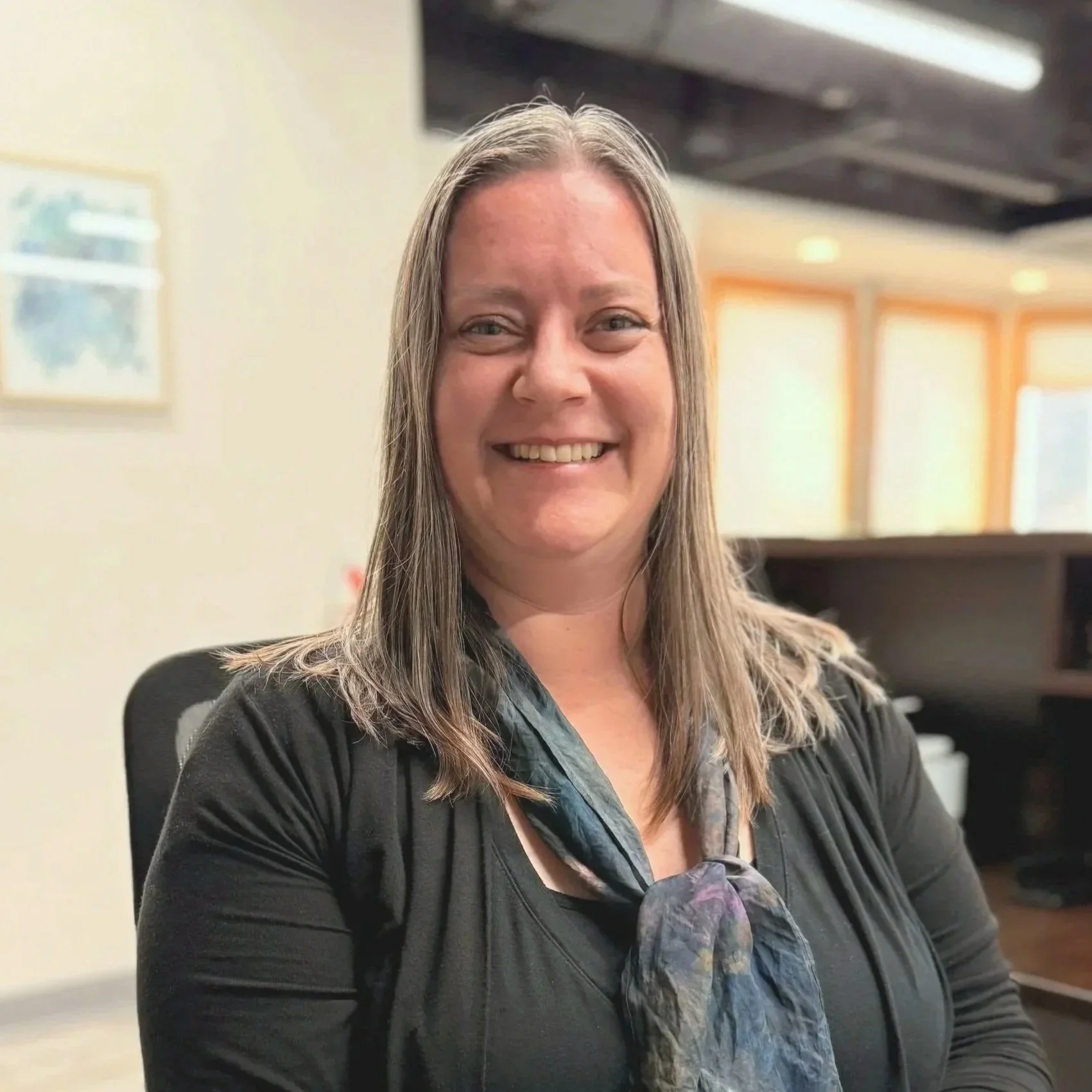 A woman with long dark hair smiling, wearing a white top, maroon cardigan, and black pants against a plain white background.