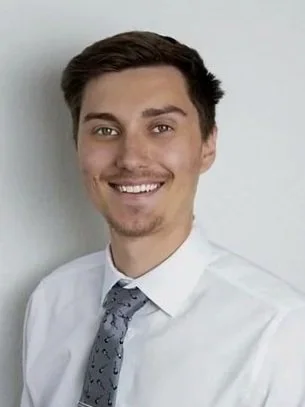 A man with short dark hair, light skin, and brown eyes is smiling slightly at the camera. He is wearing a collared shirt, with a plain blue background.
