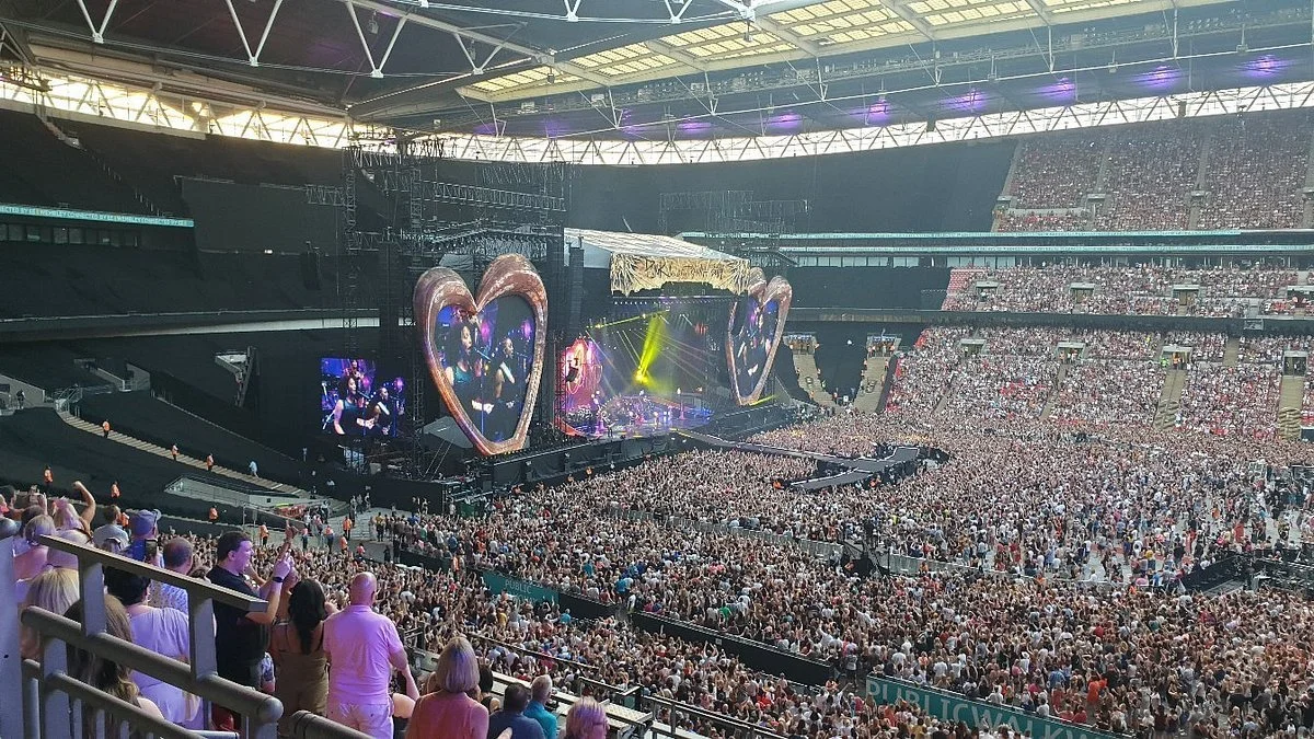 Large crowd at a concert in a stadium with a stage decorated with heart-shaped frames, and colorful lighting.