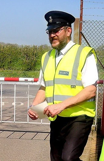 Security guard wearing a neon yellow reflective vest, a security cap, sunglasses, and holding a paper, standing outside near a gate with barbed wire and greenery in the background.