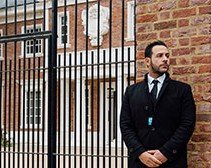 A man in a black suit and tie standing outside near a black fence, with brick and wooden buildings in the background.