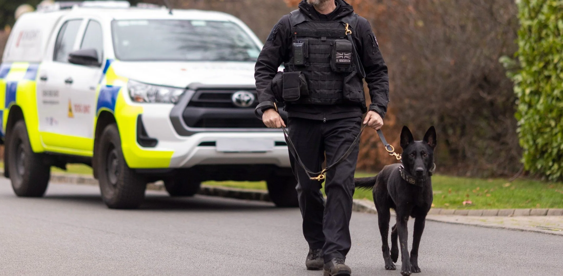 A police officer walking with a police dog on a street, with a police vehicle in the background.