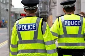 Two police officers wearing high-visibility yellow jackets and police hats, standing on a city street.