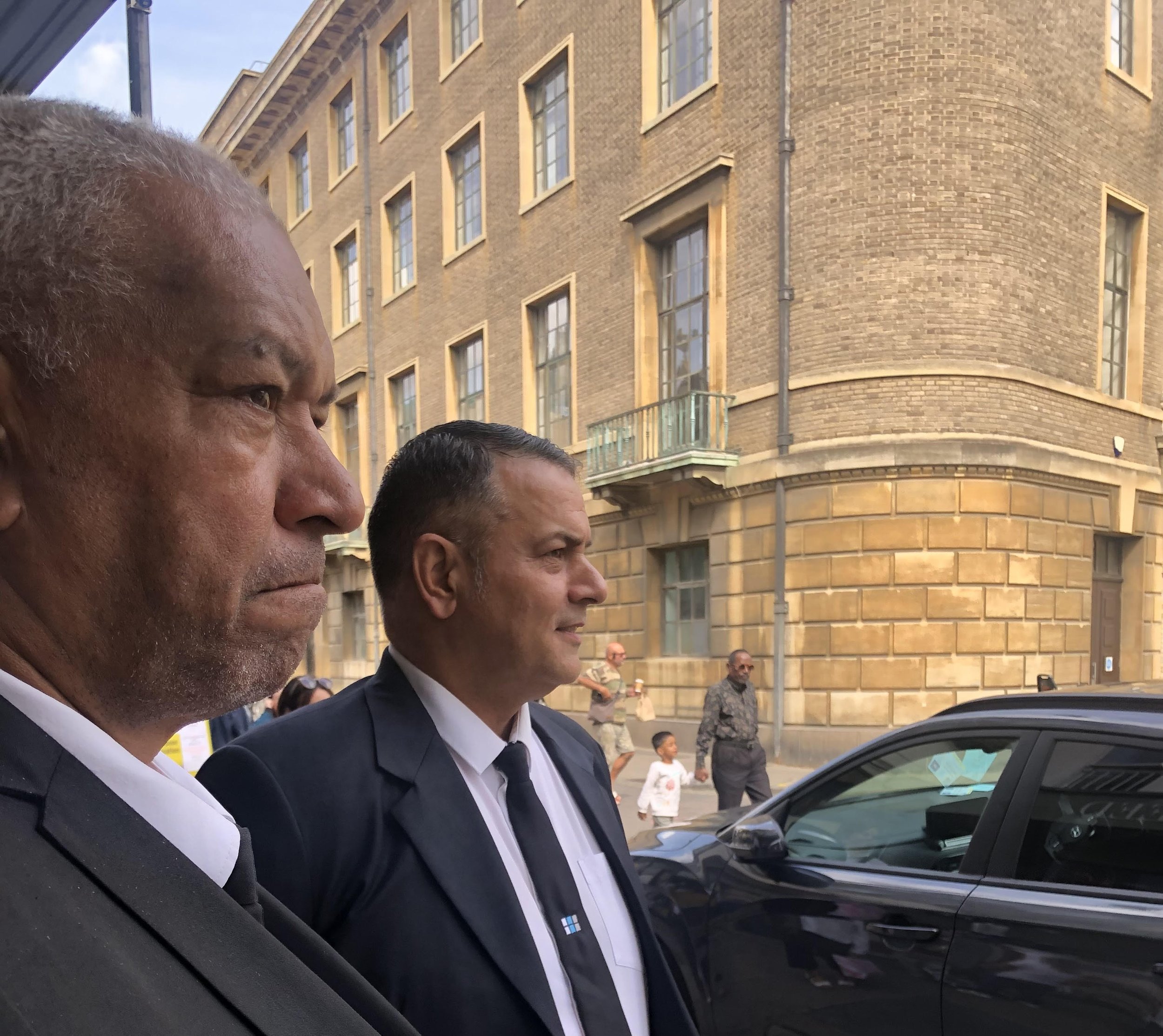 Two men in suits standing on a city street with old brick building in the background, and a black car parked nearby.