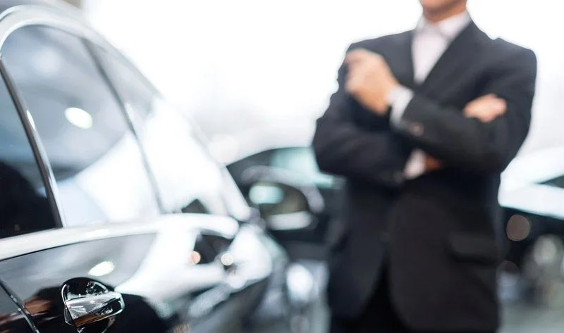 A man in a suit with arms crossed standing near cars in a showroom or dealership.