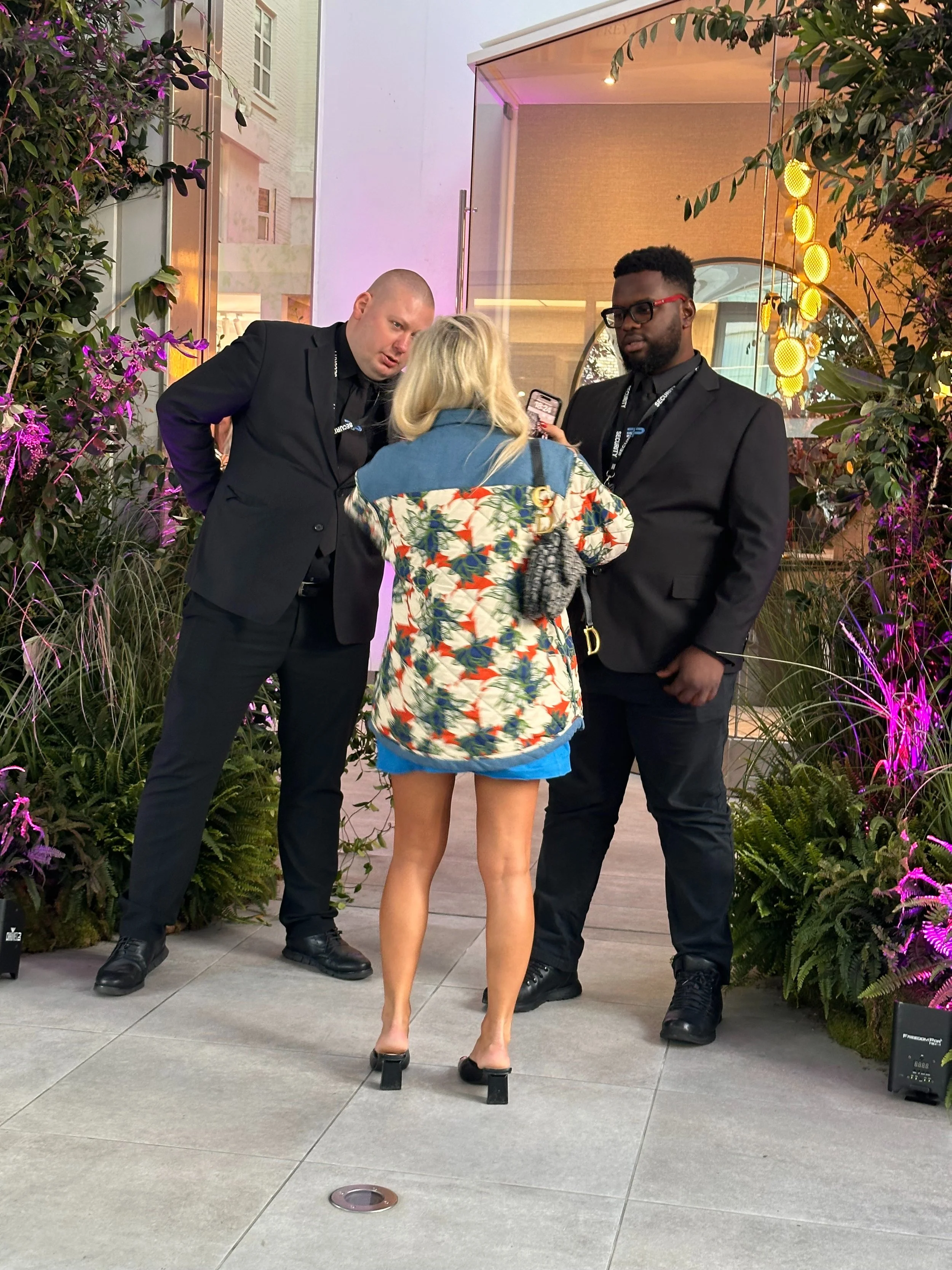 A woman in a floral jacket and high-heeled shoes is talking to two men in black suits outside an event venue, with greenery and colorful lights in the background.
