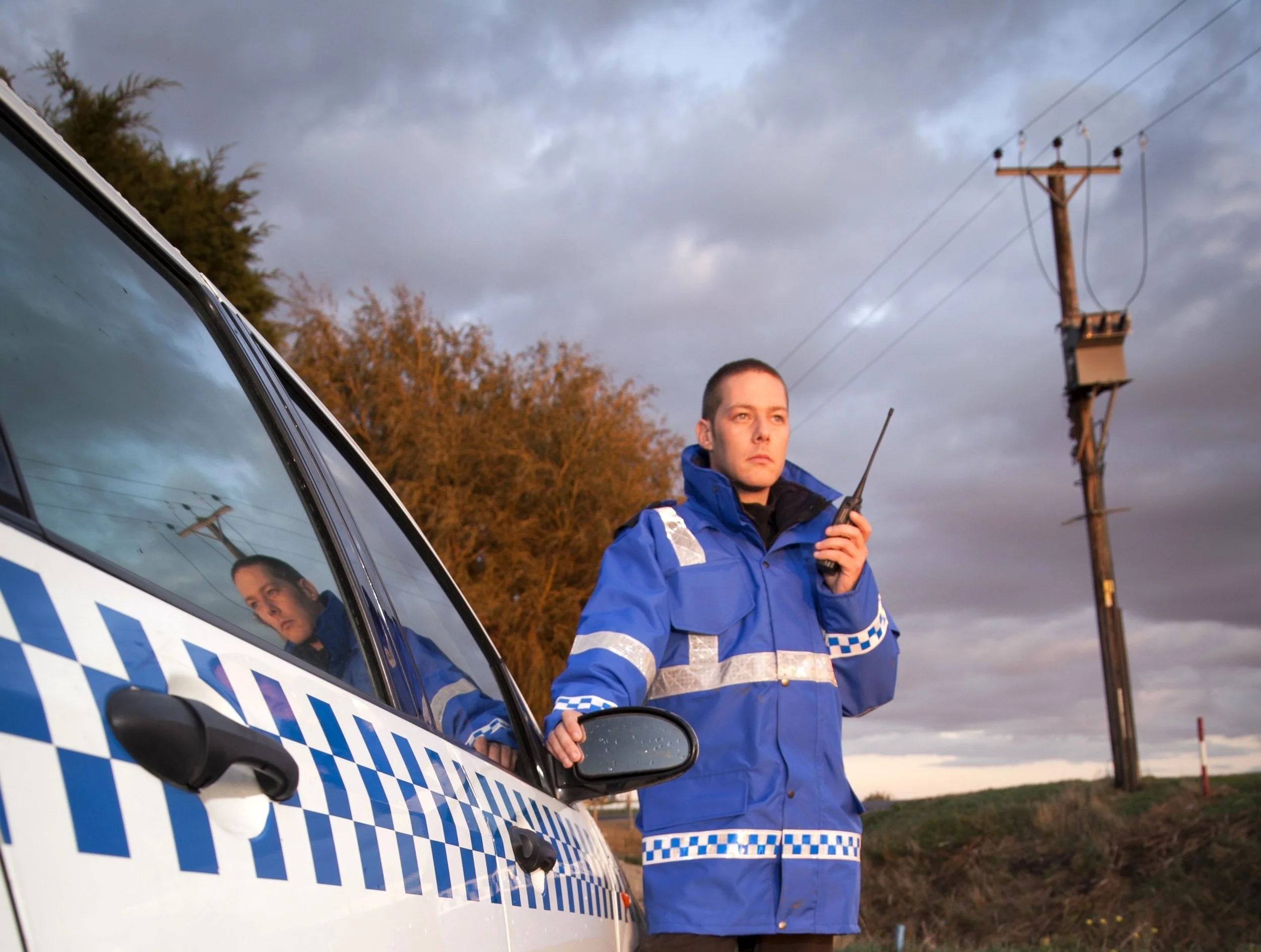 A female police officer standing outside a police car with a walkie-talkie, during sunset with clouds in the sky, and power lines and trees in the background.