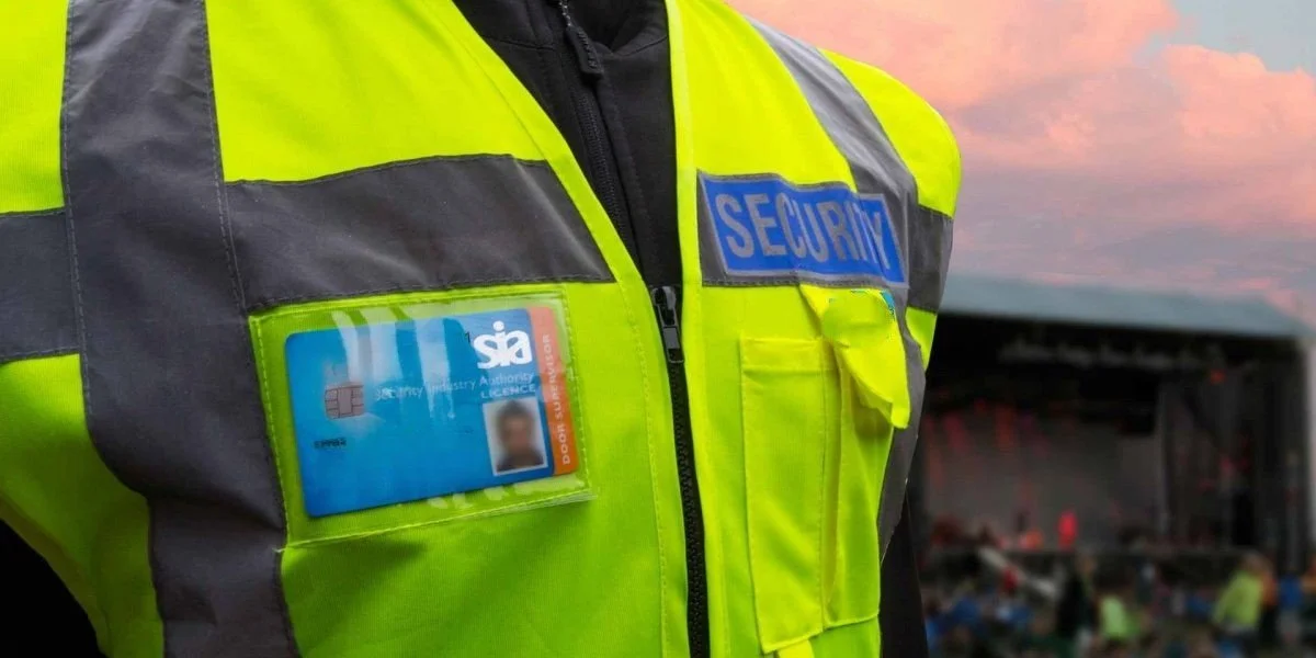 Close-up of a security guard's bright yellow and gray uniform with a blue 'SECURE' patch and an ID badge, against a background of an outdoor stage at sunset.