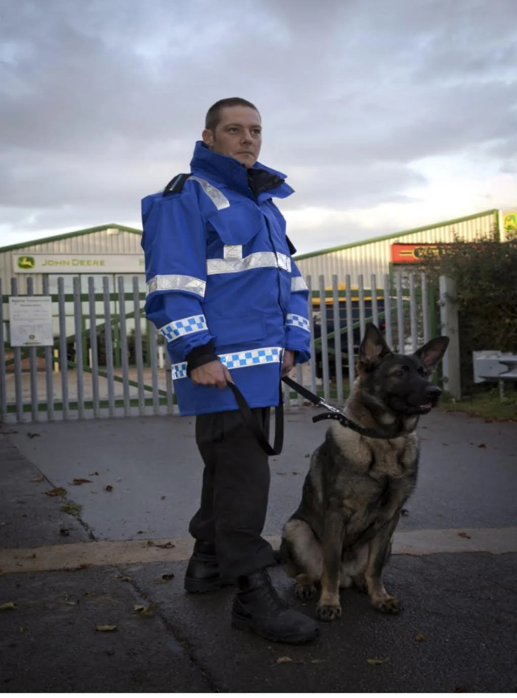 A police officer in a blue uniform with reflective stripes standing outdoors with a German Shepherd police dog. The background shows a fence and a John Deere sign, under a cloudy sky.