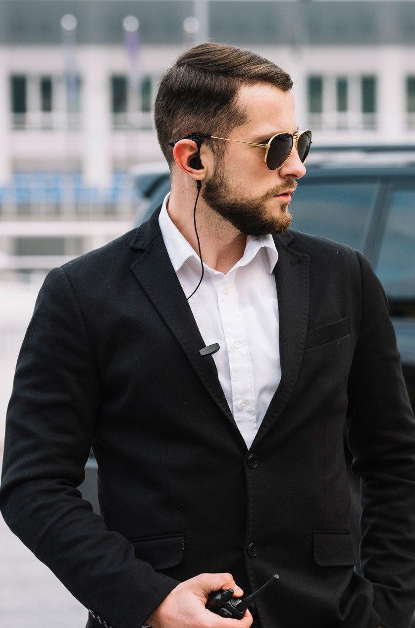 A man wearing sunglasses and a black blazer, holding a walkie-talkie, standing outdoors near a vehicle.