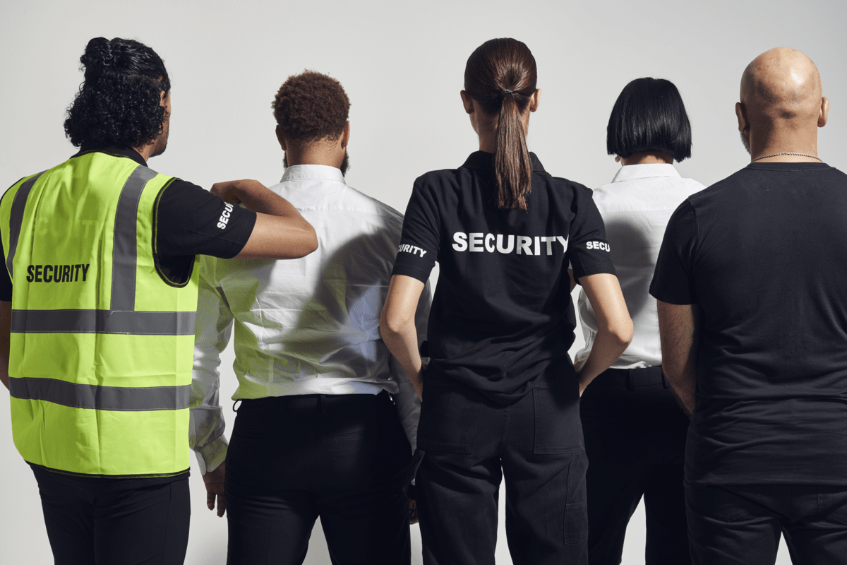 Group of security personnel, including two women and three men, standing with their backs facing the camera against a plain white wall.