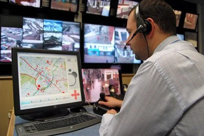 Man working at a control station with multiple monitors, including a computer displaying a map and security footage.