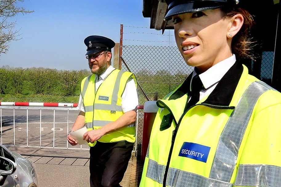 Two security guards in high-visibility yellow jackets standing outdoors near a gate, with a man in the background, open car door, and a chain-link fence.
