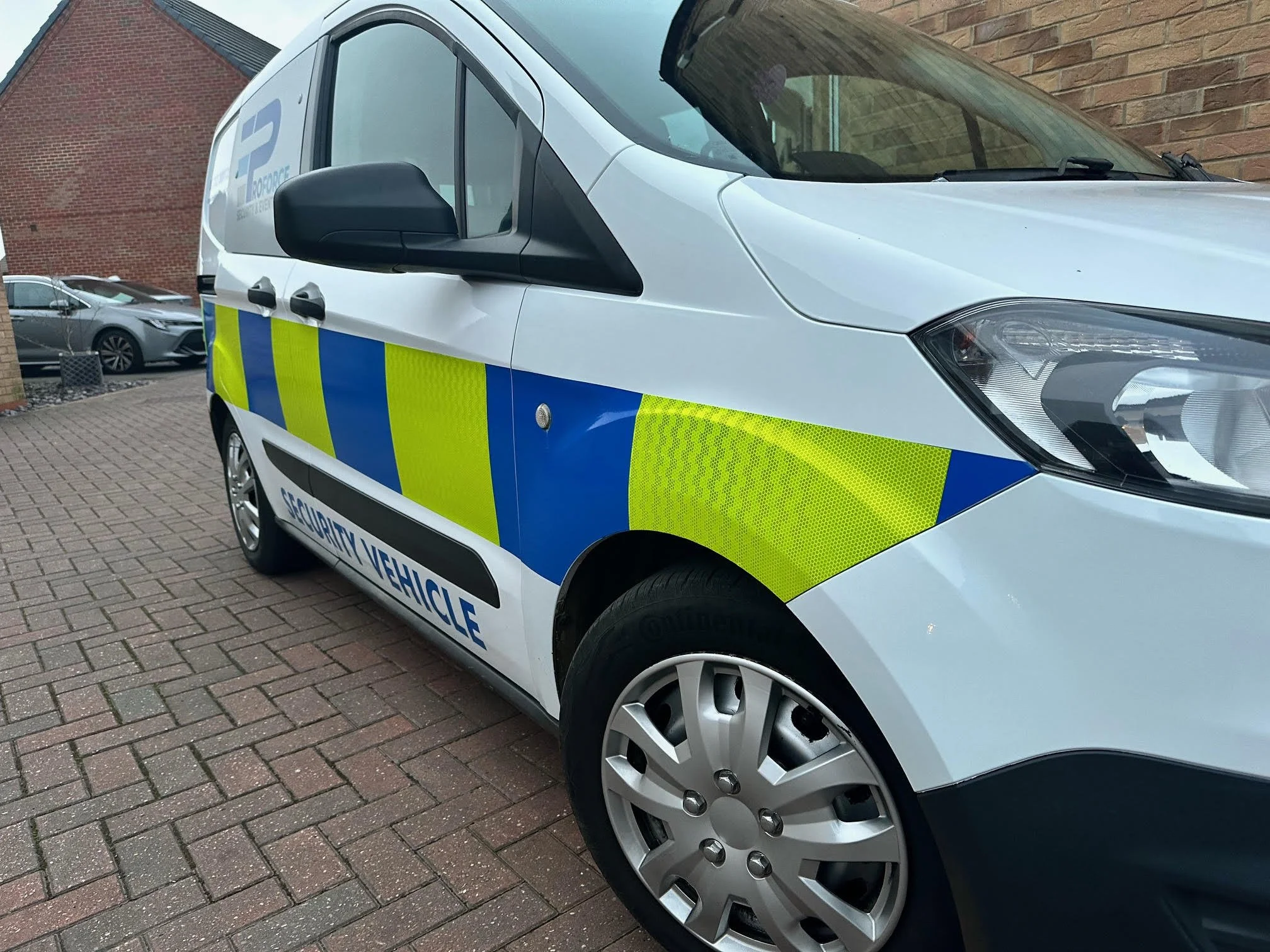 A white security vehicle with blue and yellow reflective stripes parked on a brick pavement next to a brick wall and other cars in the background.