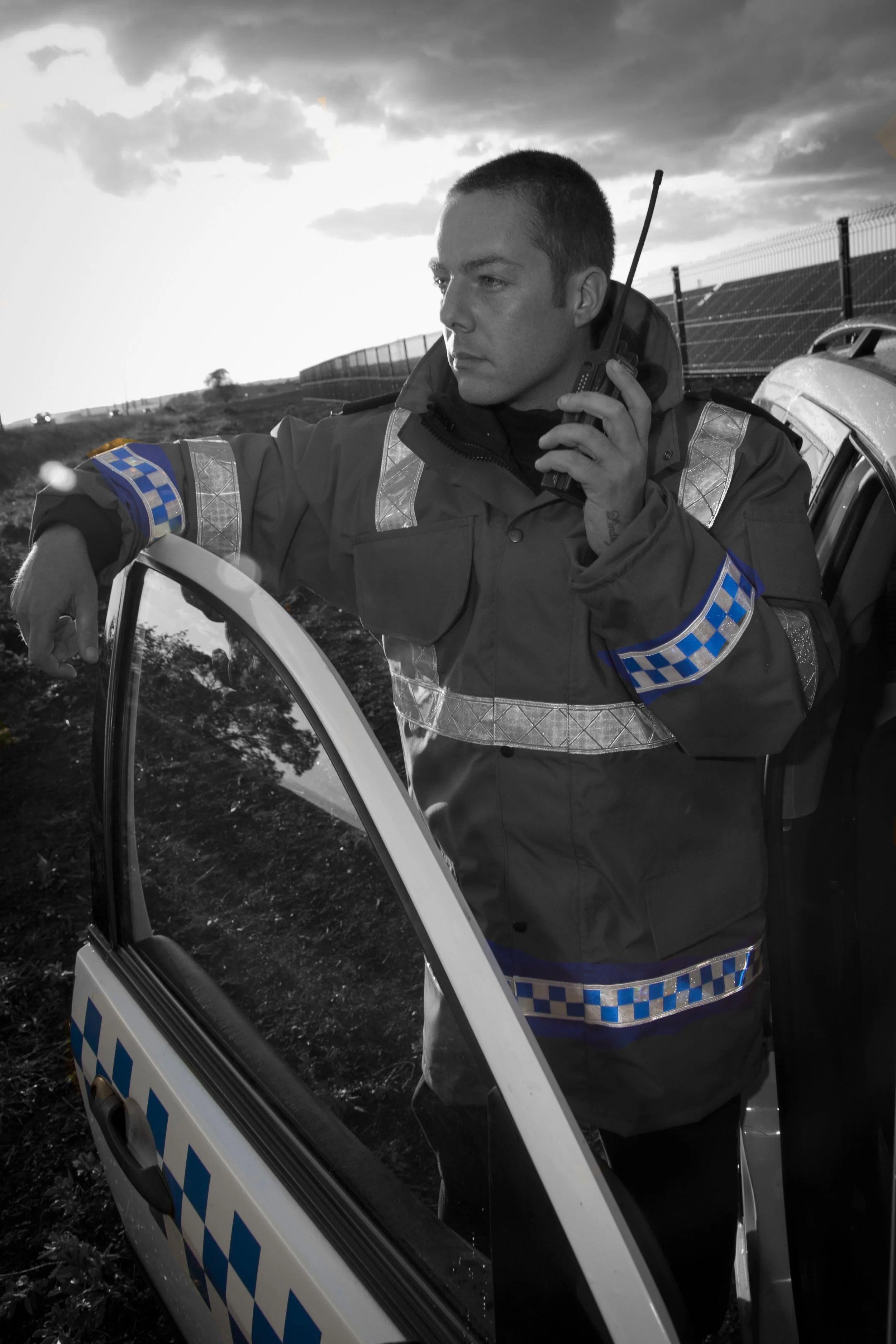 A police officer dressed in a uniform with reflective stripes is standing next to a police car, speaking on a radio, with a fence and cloudy sky in the background.