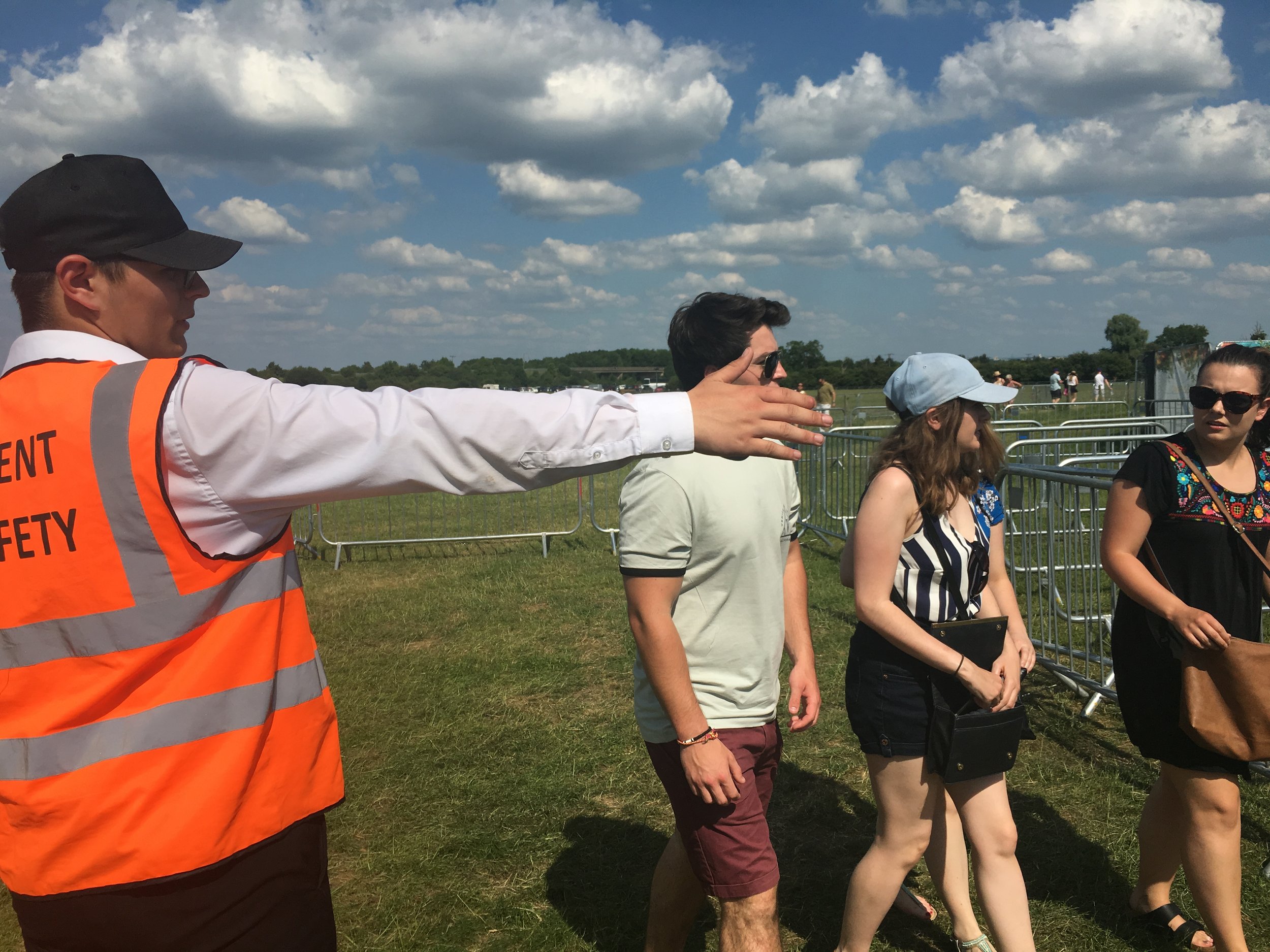 A man in an orange safety vest gesturing with his arm extended as several people walk past him outdoors on a sunny day with a blue sky and scattered clouds.