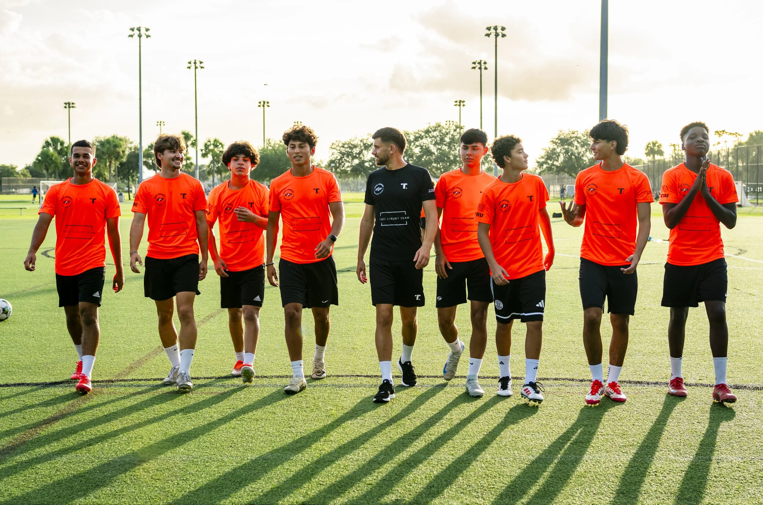 A group of young male soccer players in orange jerseys walking on a soccer field during daytime.