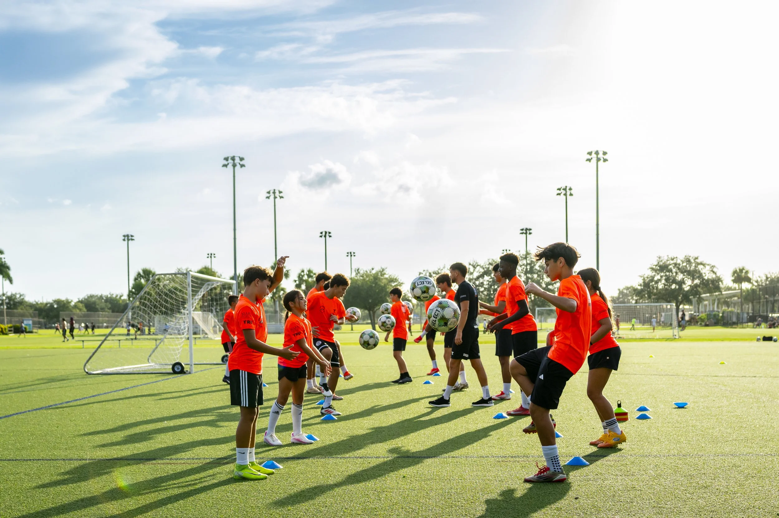 Children in bright orange shirts practicing soccer drills on a sunny field with goals and trees in the background.