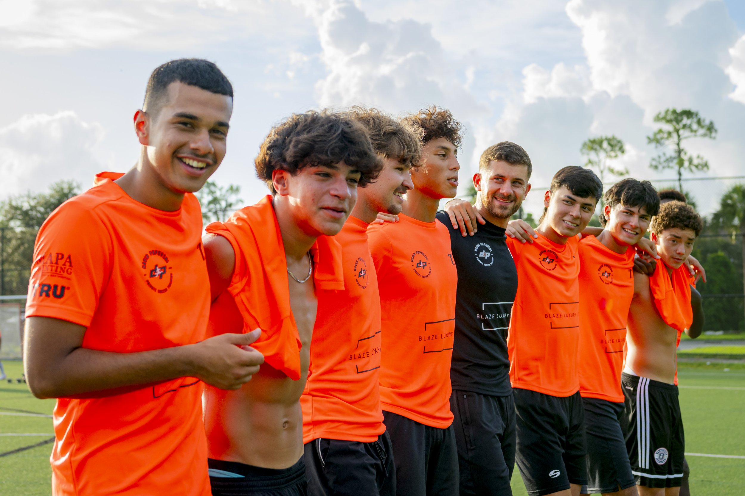 A group of eight young male soccer players standing side by side outdoors on a soccer field, wearing orange jerseys and black shorts, with some having their arms around each other's shoulders, smiling for the photo against a background of trees, grass, and a partly cloudy sky.