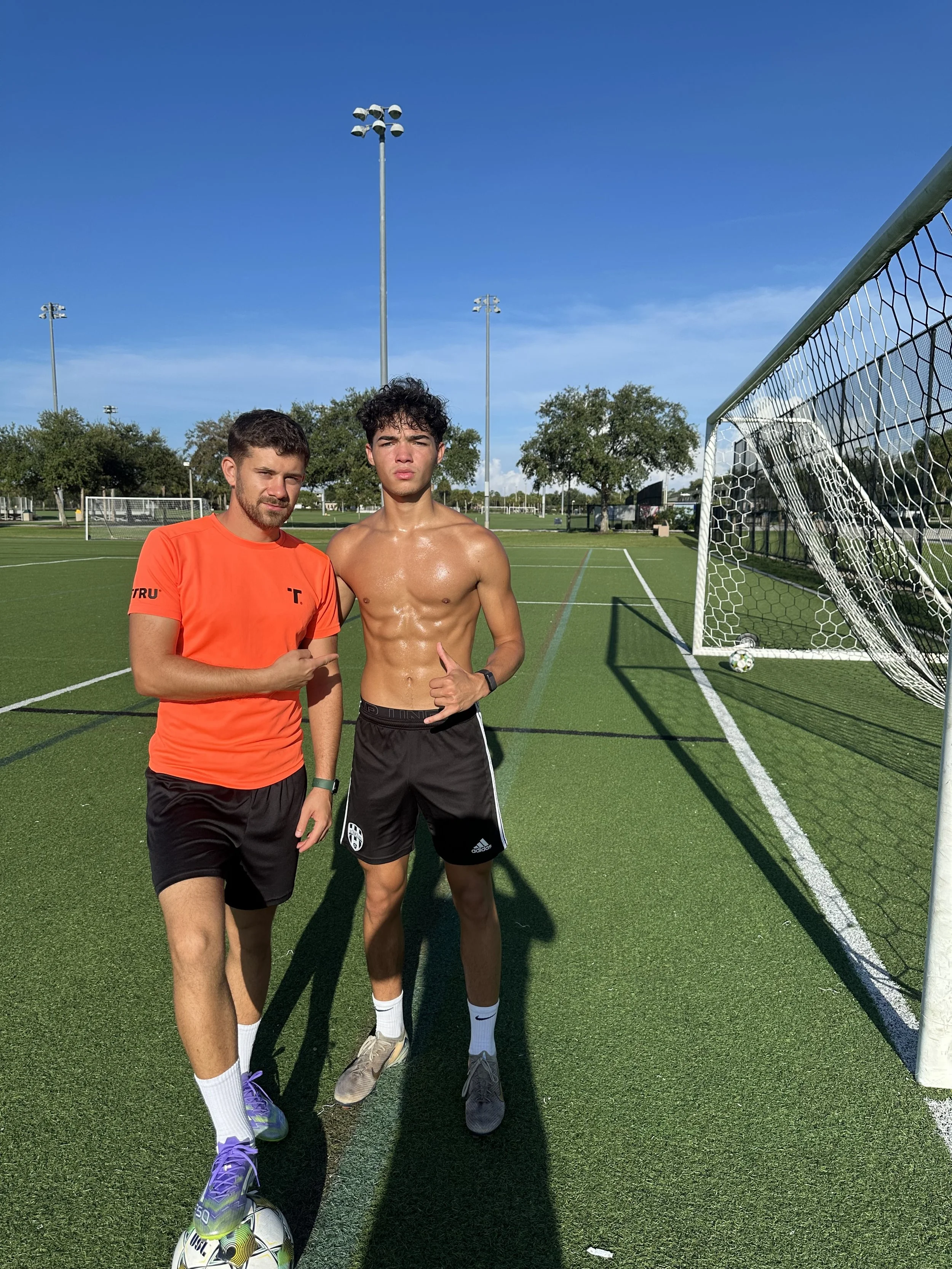 Two male soccer players standing on a soccer field near a goalpost, one shirtless and the other in an orange shirt, both with athletic builds, under a bright blue sky, with soccer balls visible on the ground.