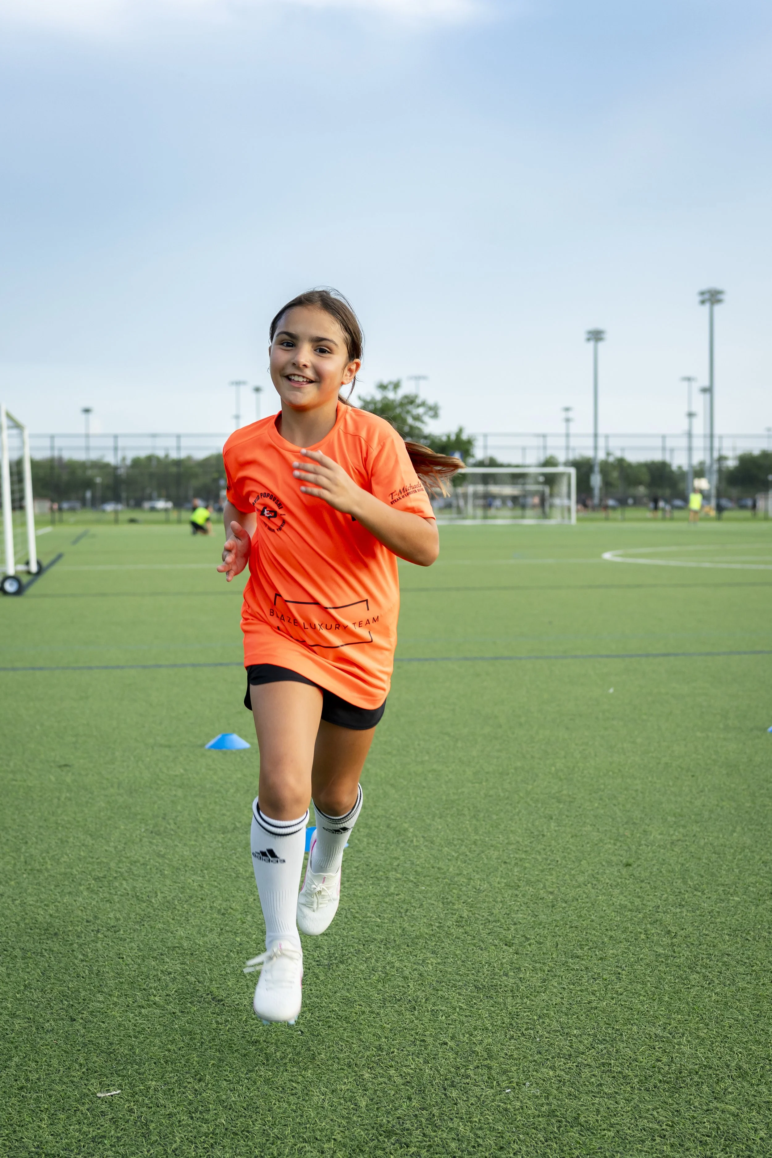 A young girl with dark hair and a bright smile running on a soccer field in an orange sports jersey and white socks with black stripes, surrounded by soccer goals and cones.