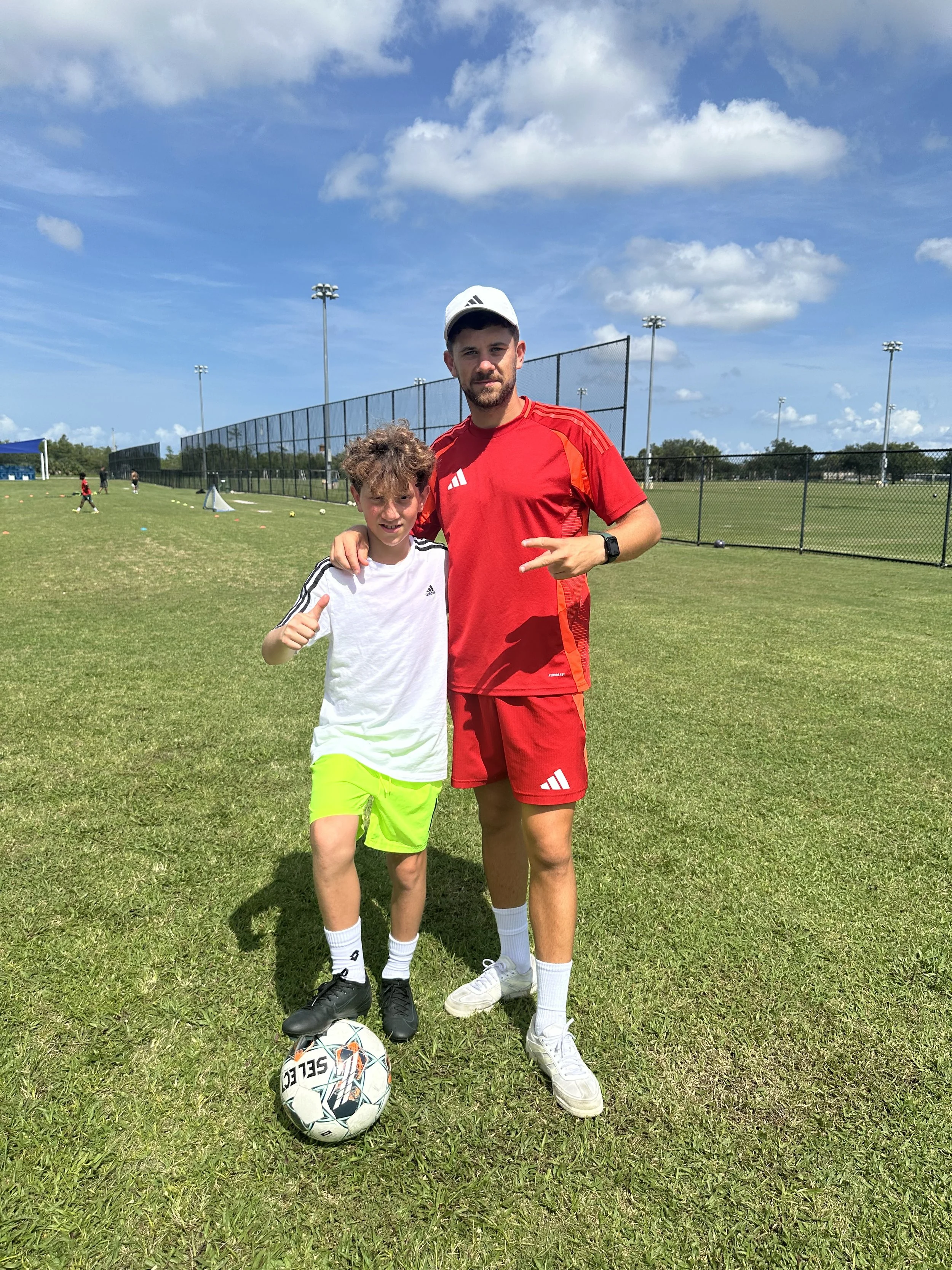 A young boy and an adult man standing on a grassy soccer field, with the boy's arm around the man's shoulder. The boy is giving a thumbs-up and has a soccer ball under one foot. The man is making a peace sign with his fingers. Both are dressed in athletic clothing, and there are other players in the background.