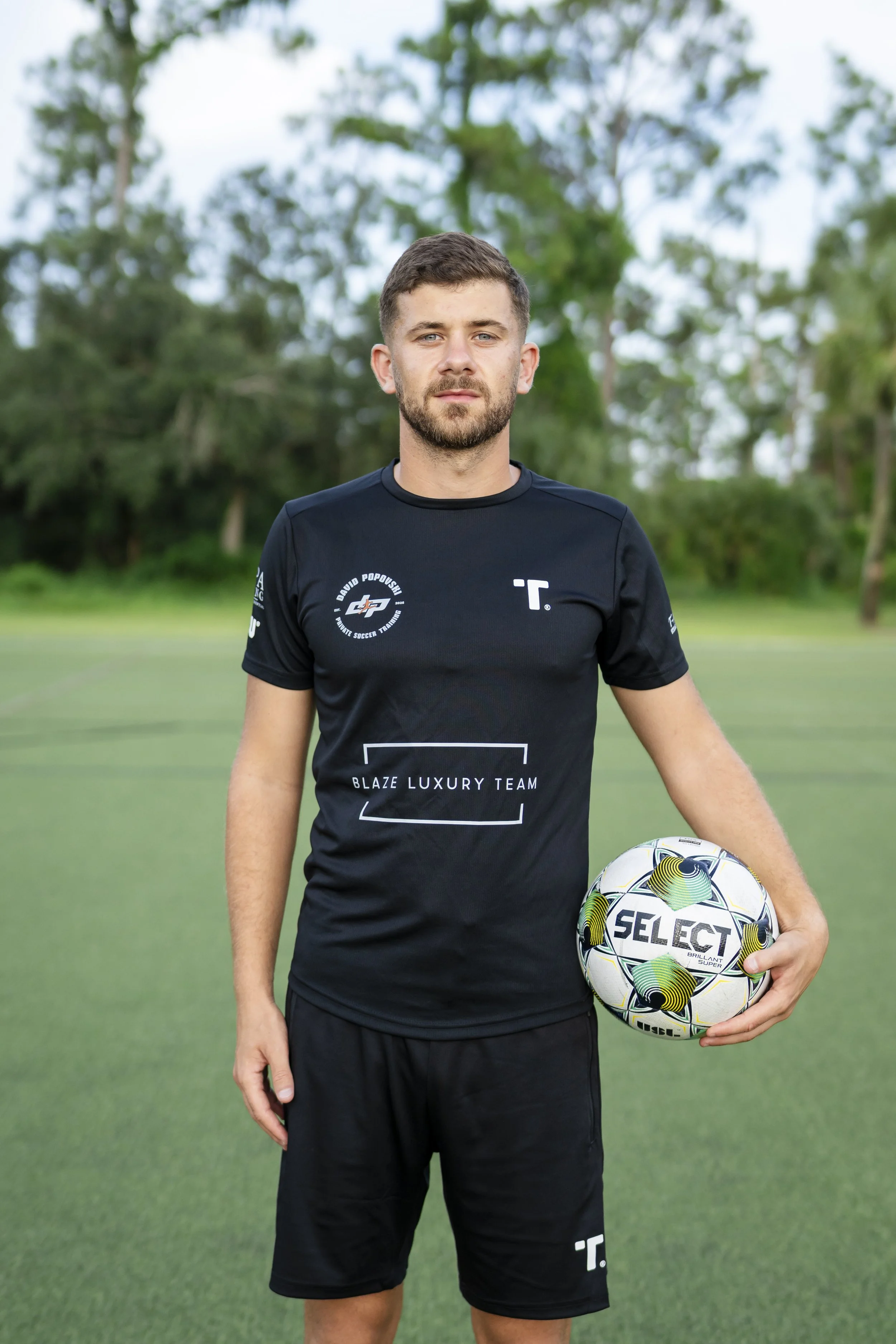 A young man in a black soccer uniform holding a soccer ball on a grass field with trees in the background.