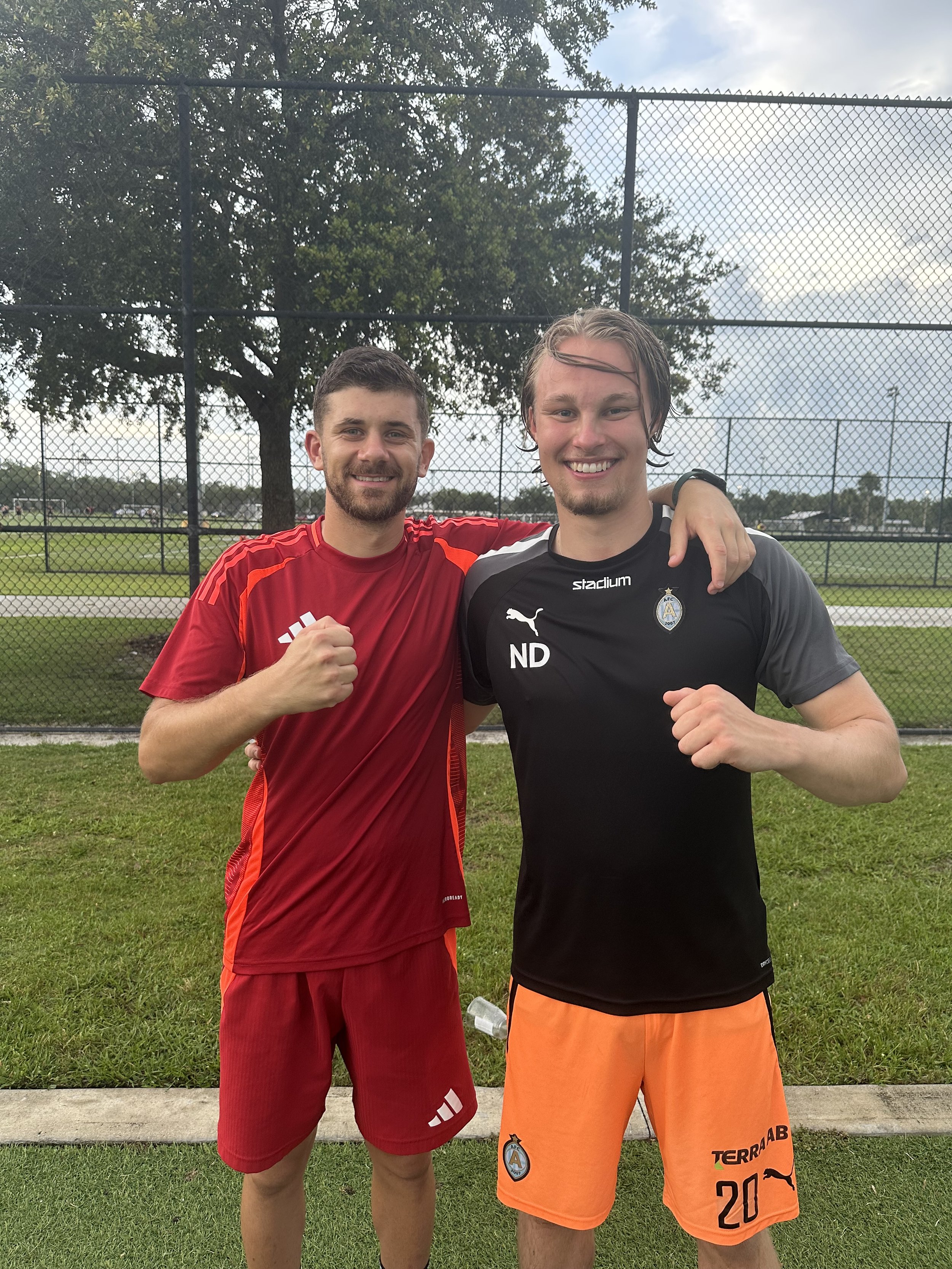 Two young men in athletic shirts and shorts are standing on grass with a chain-link fence and trees in the background. They are smiling and posing with fists raised in a friendly manner.