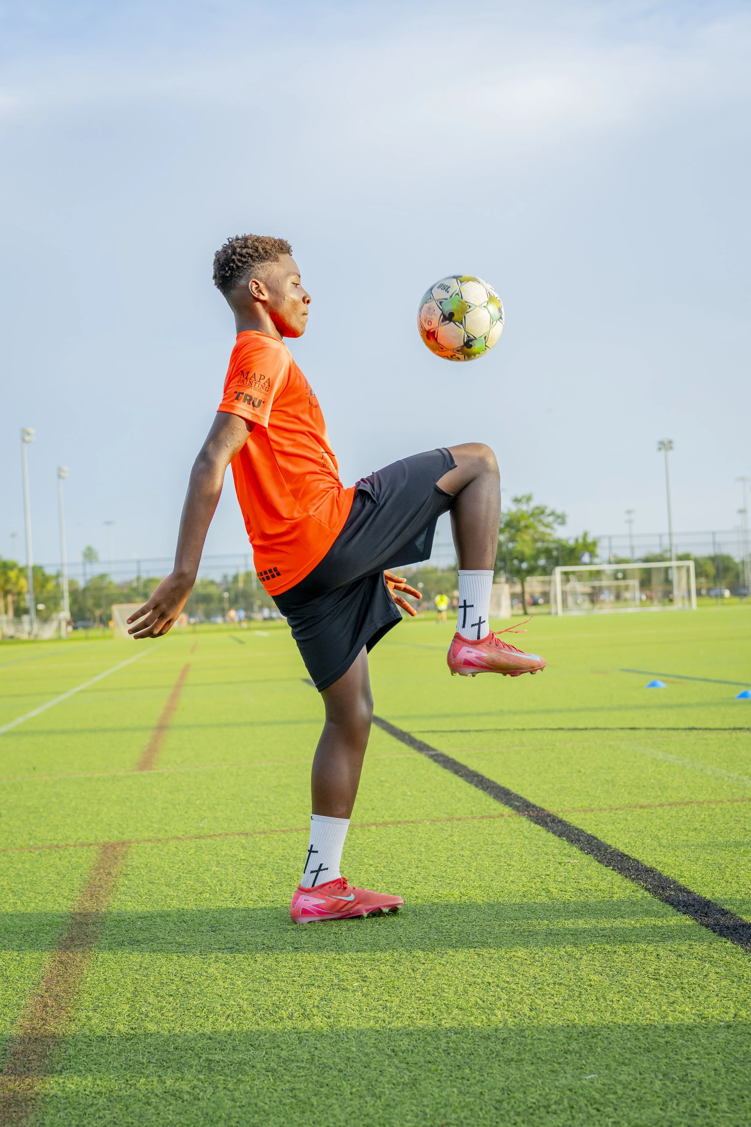 Young male soccer player in bright orange jersey and black shorts practicing on a green field, balancing a soccer ball on his knee during daytime.
